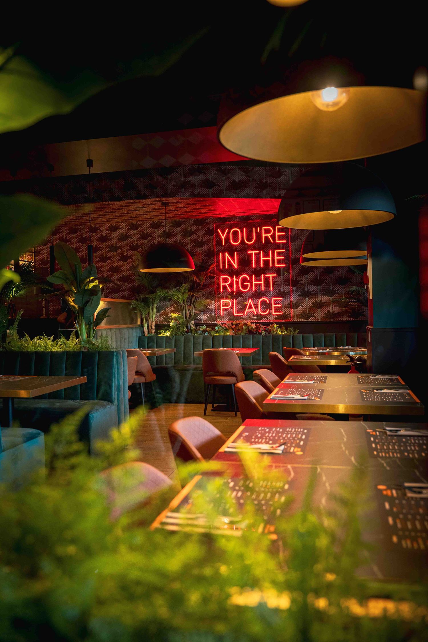 Stylish restaurant interior with red neon sign reading You're In The Right Place and lush greenery.