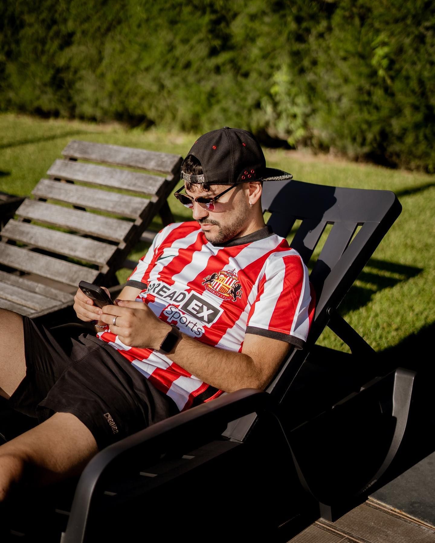 Man in striped soccer jersey relaxing on a lounge chair outdoors using his smartphone.
