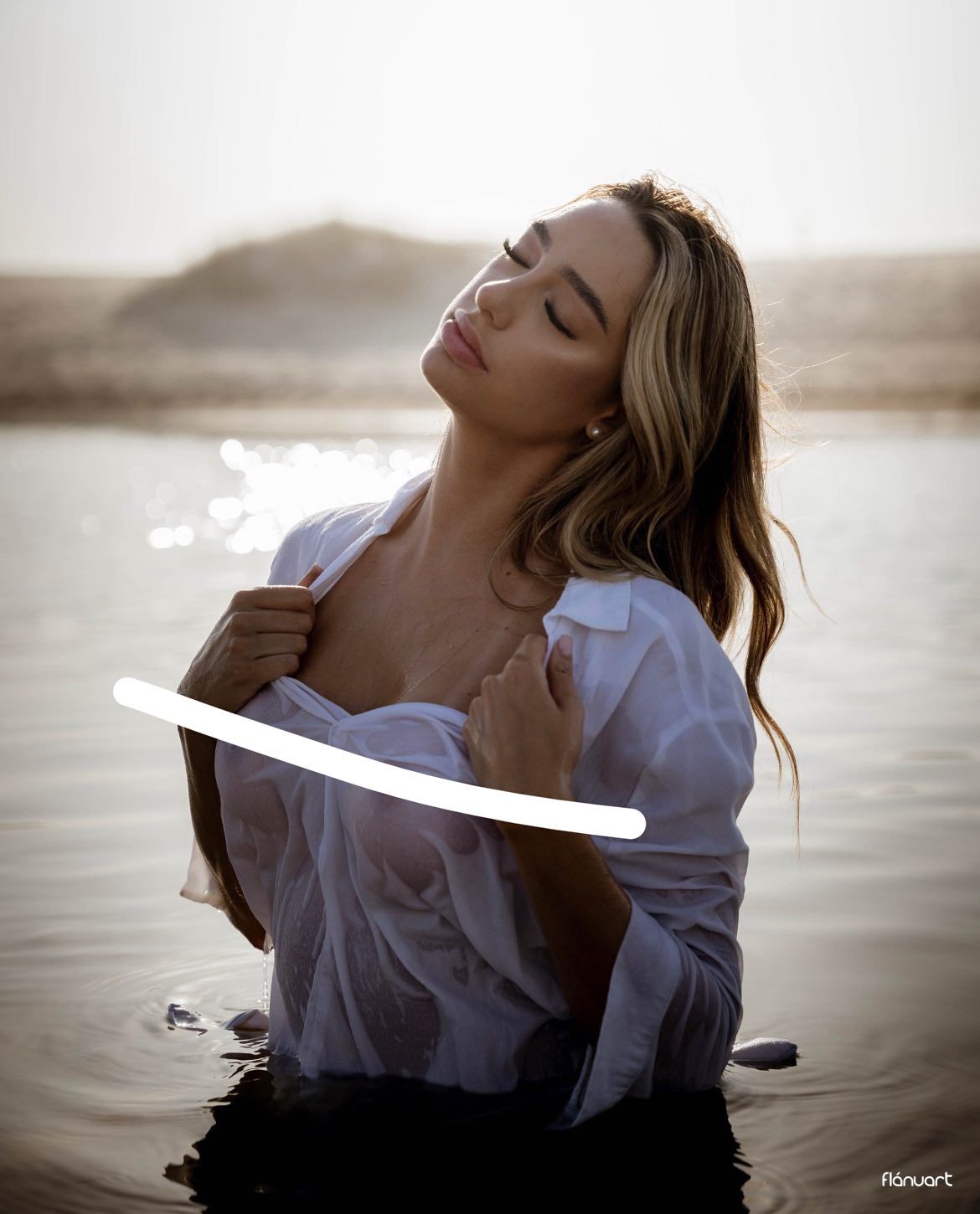 Woman in wet white open shirt standing in shallow water during misty golden hour light.