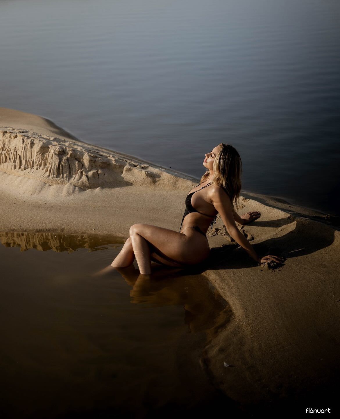 Woman in black bikini sitting on sandy shore beside calm water with dramatic sand dunes in background.