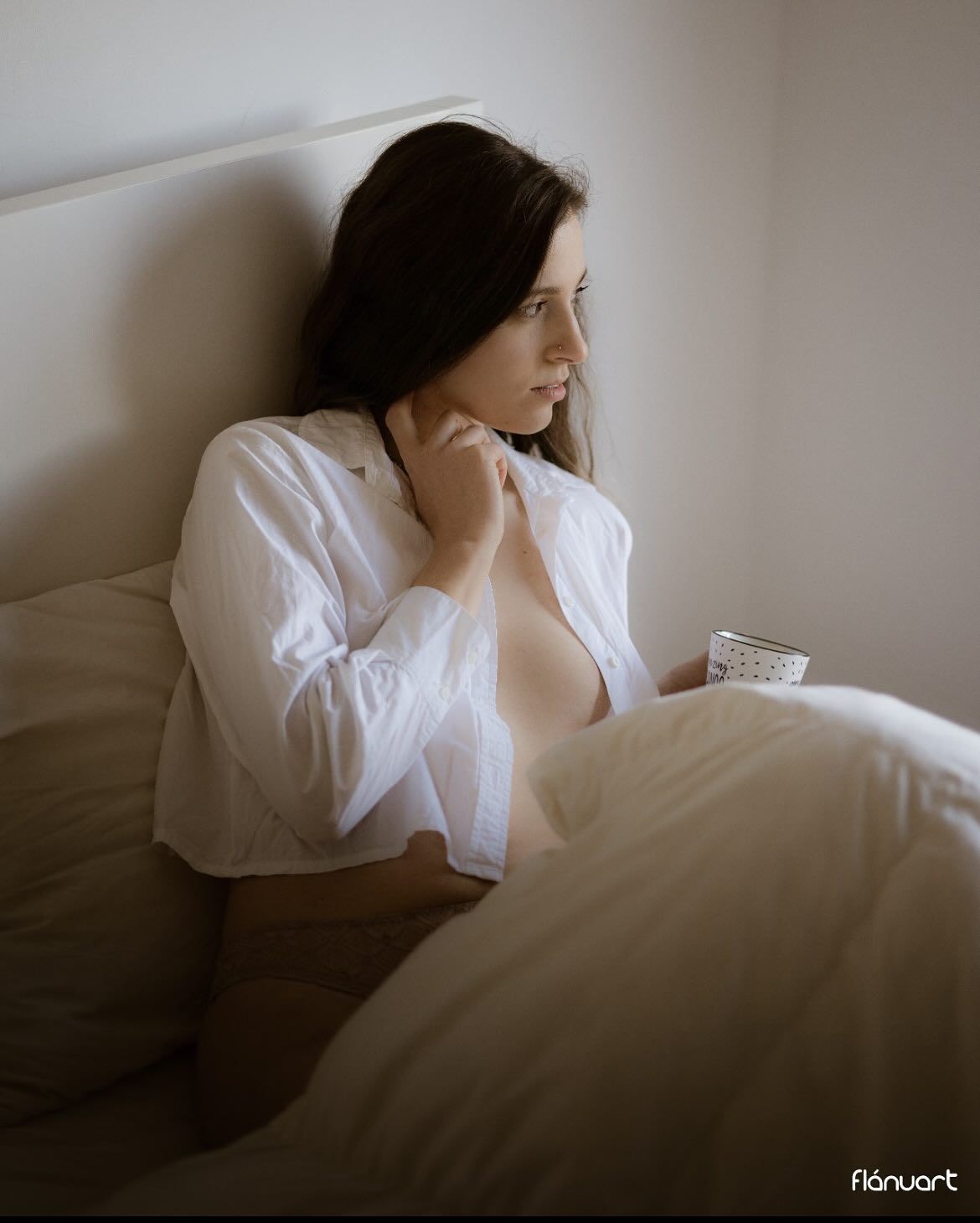 Brunette woman in open white shirt sitting in bed holding coffee cup in soft light.