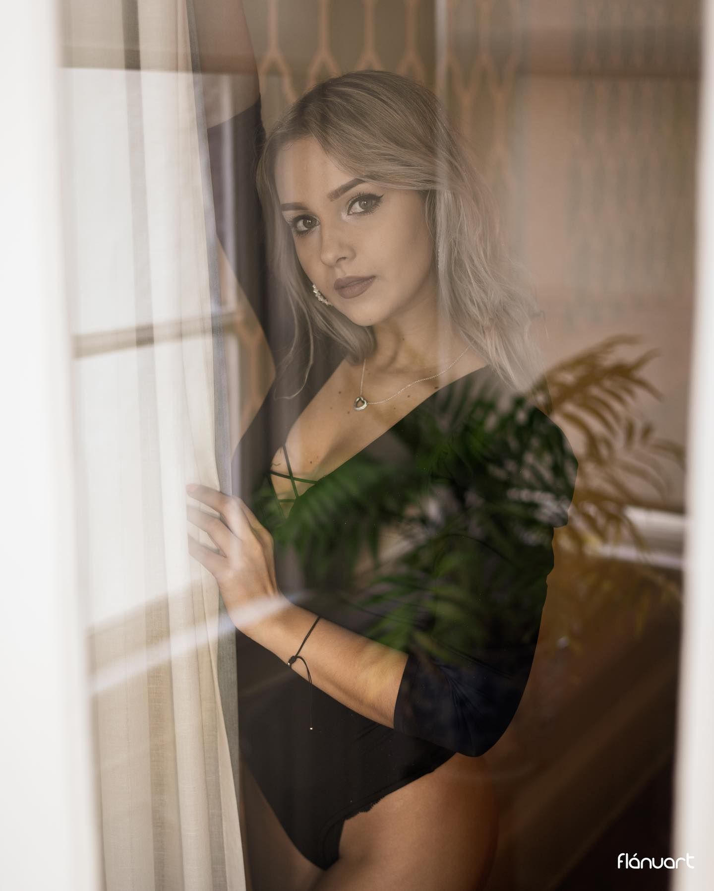 Blonde woman in black bodysuit gazing through sheer curtains by a window.