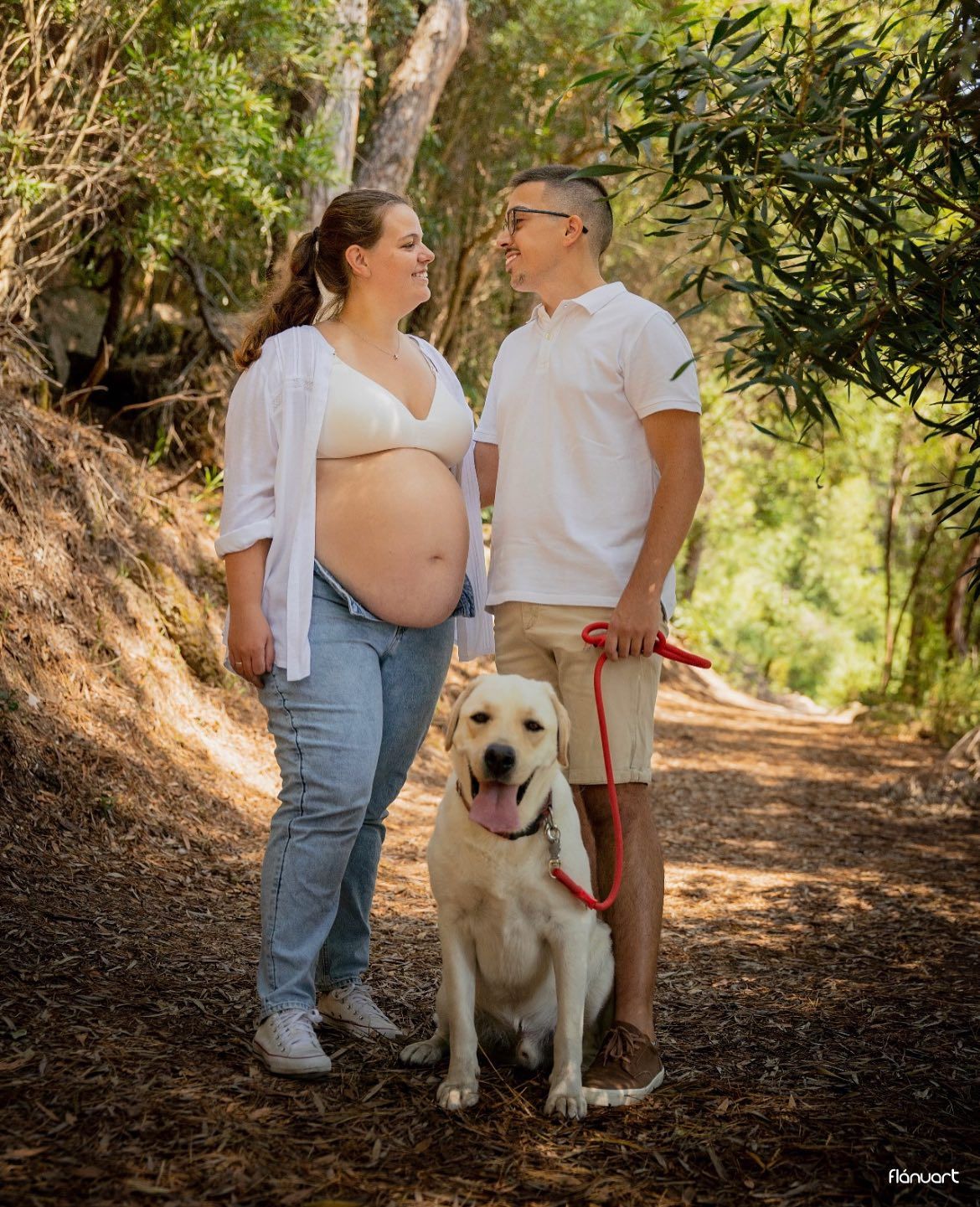An expectant couple smiles at each other on a forest trail with their happy yellow Labrador dog.
