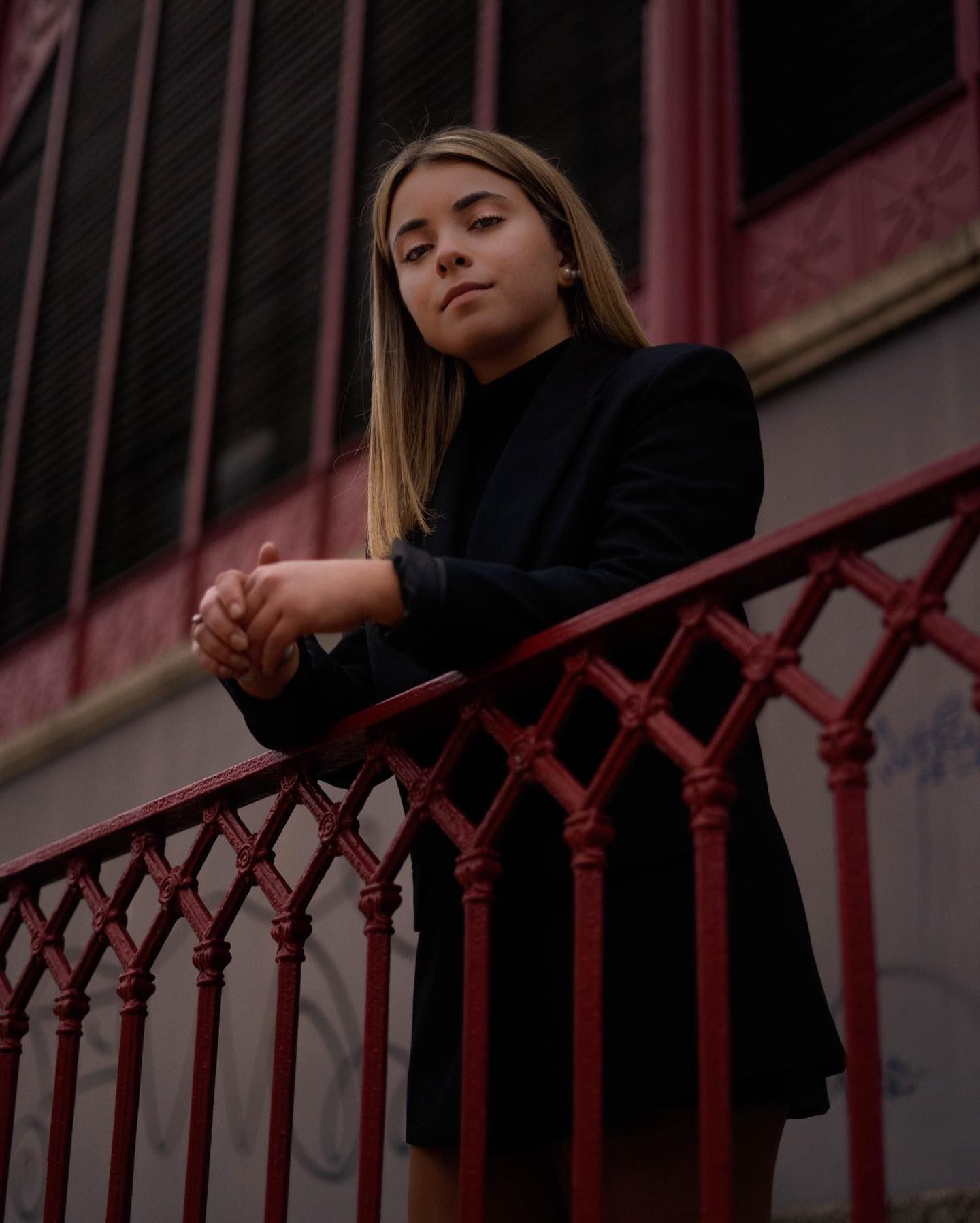 Young blonde woman in a black blazer leaning on a red ornate railing outdoors.