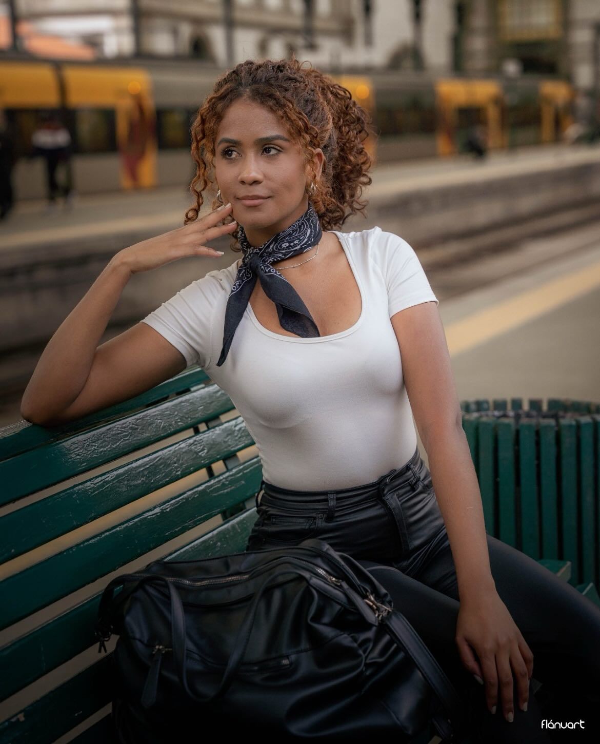 Young curly-haired woman in white top and black skirt sitting on a bench at a train station platform.
