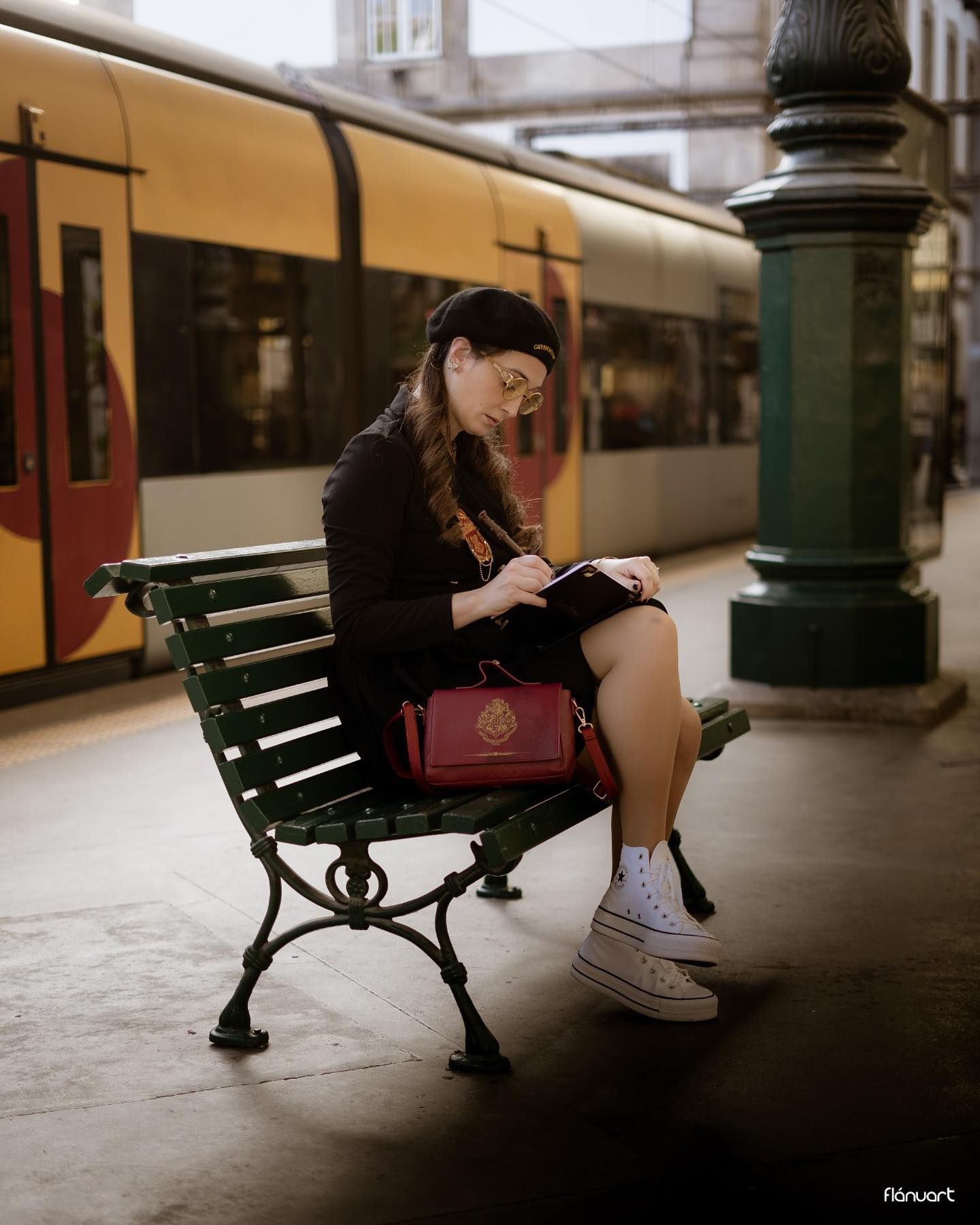 Young woman in black outfit sitting on bench reading at classic European train station platform.