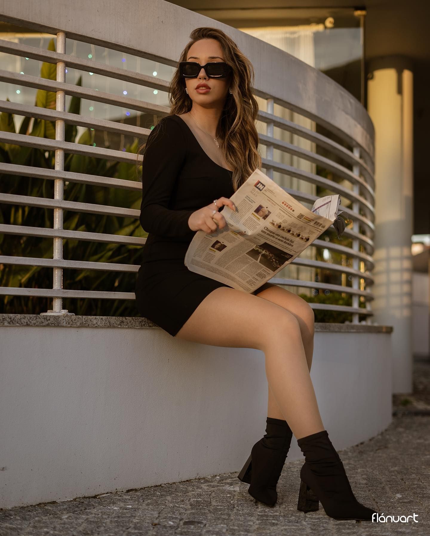 Woman in black mini dress and boots sits on ledge reading newspaper wearing sunglasses.