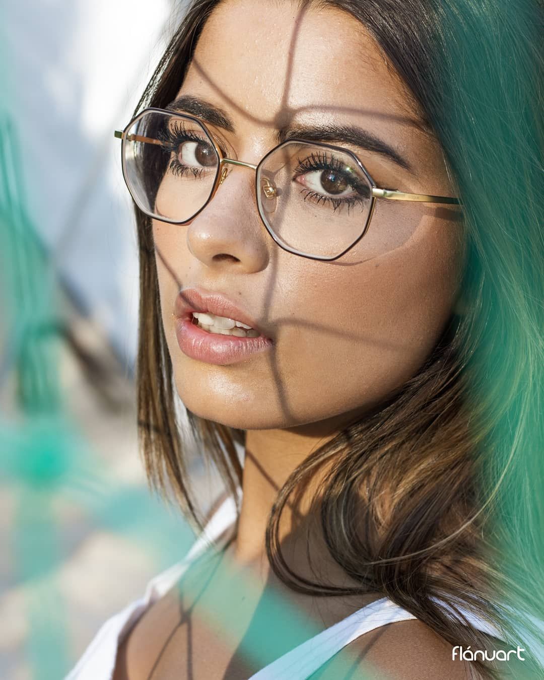 Close-up portrait of a young woman wearing round gold-framed glasses with teal hair highlights.