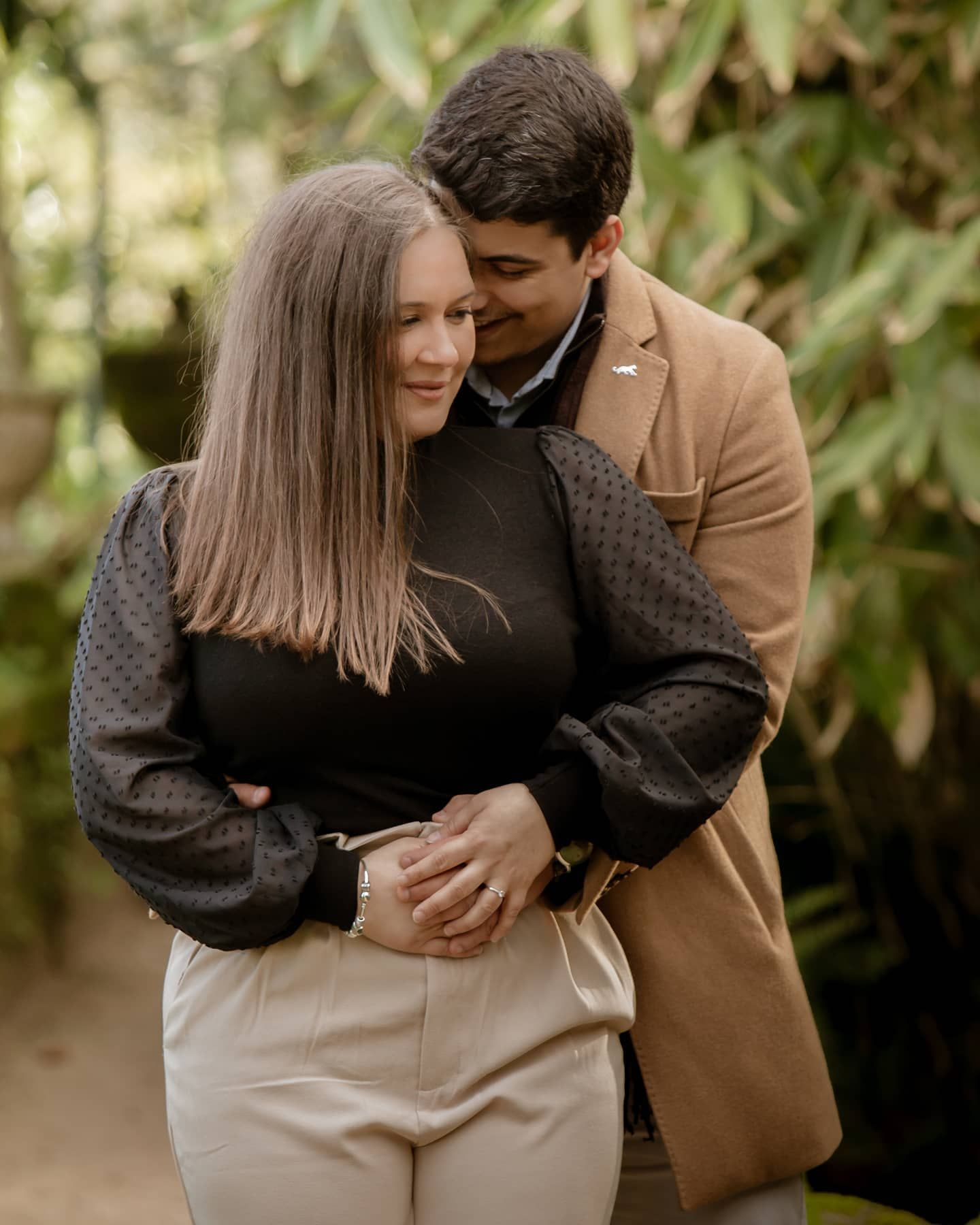 Couple embracing tenderly in a green garden, woman in black top, man in camel blazer holding her close.