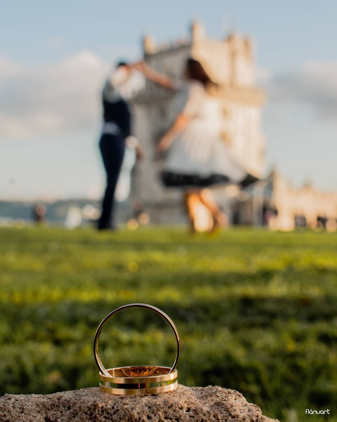 Gold wedding rings on a rock with a couple dancing near Belem Tower in Lisbon background.