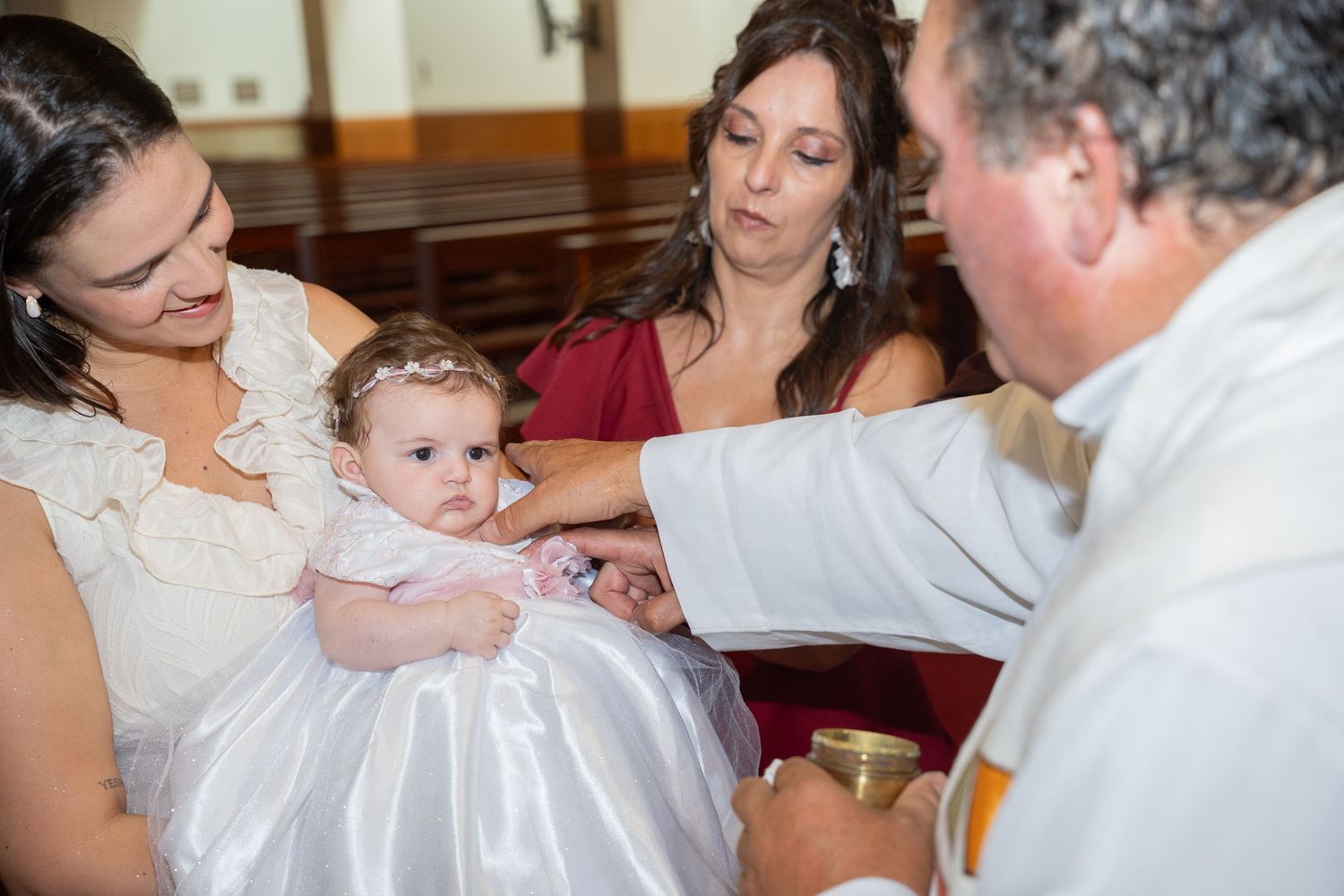 A priest baptizing a baby girl in a white dress, held by her mother, during a church christening ceremony.