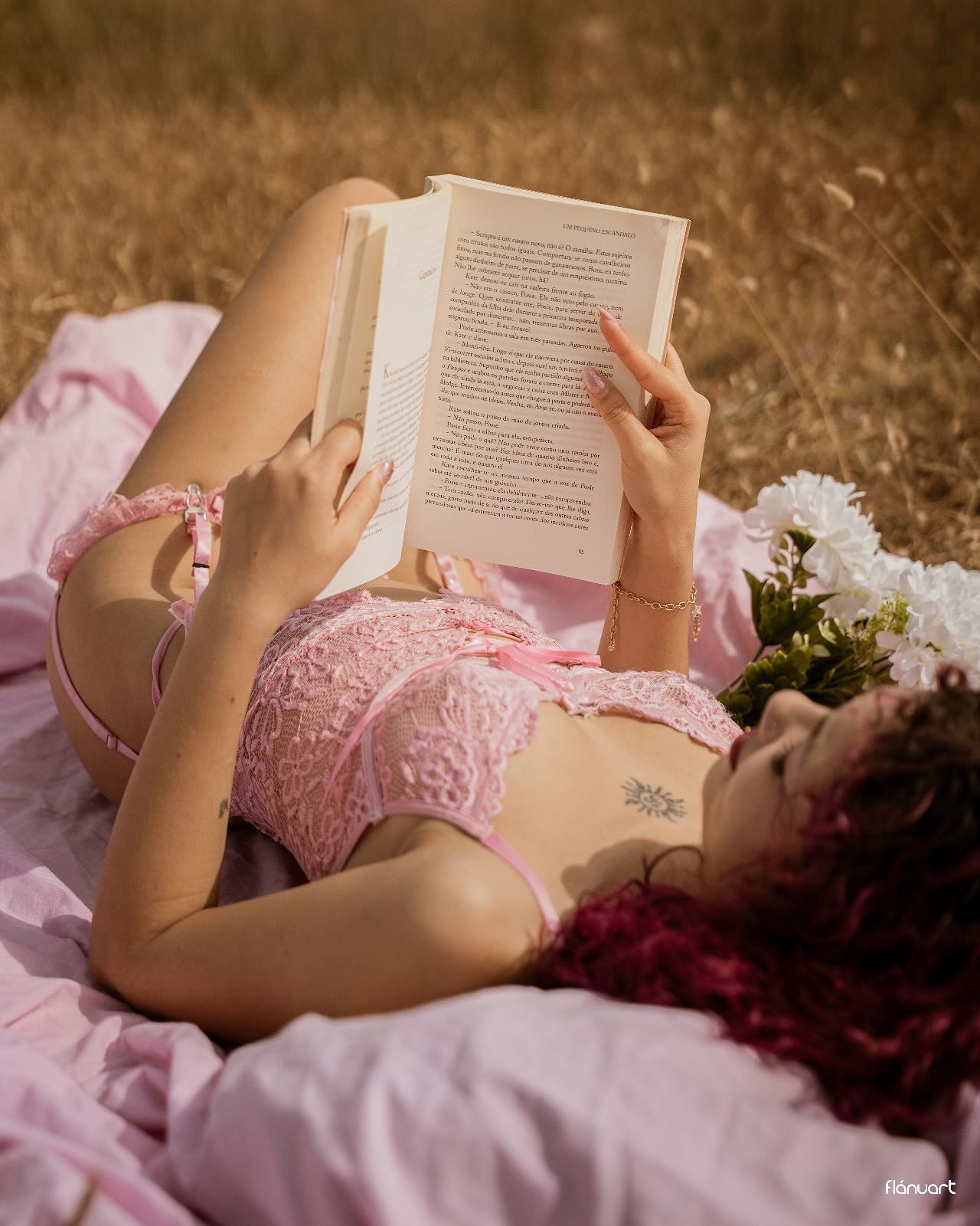 Woman in pink lace lingerie lying in golden field reading a book with white flowers nearby.