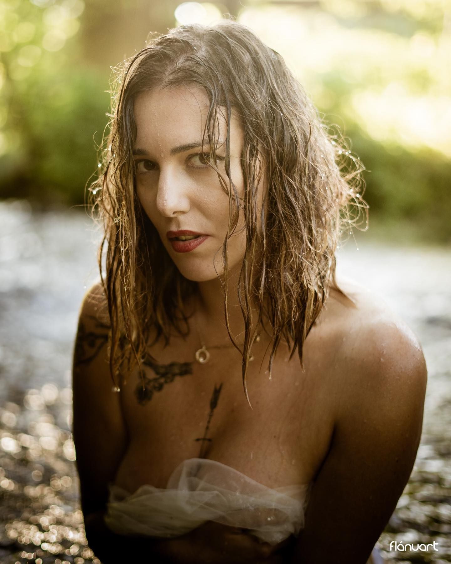 Portrait of wet-haired woman with tattoos in natural water setting with dappled sunlight.