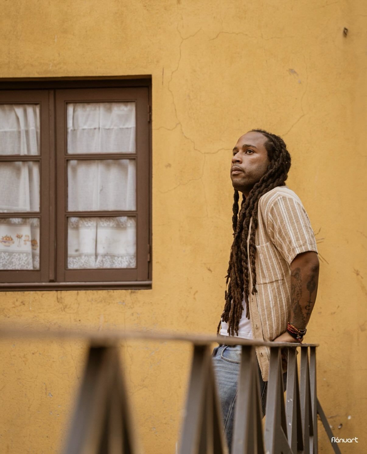 Man with long dreadlocks in striped shirt leaning on a railing against a yellow wall outdoors.