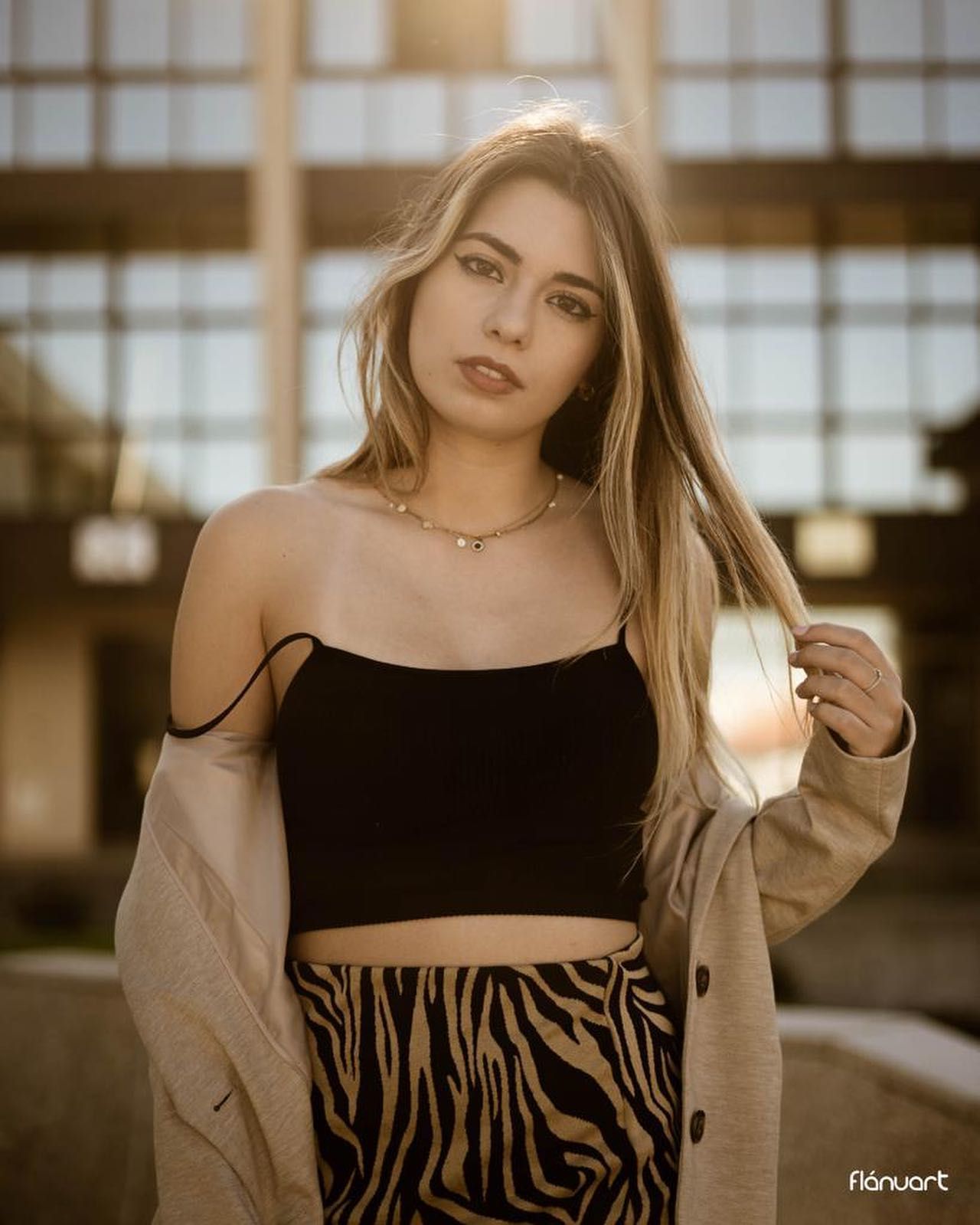 Blonde woman in black crop top, zebra print skirt, and beige coat posing in front of a blurred building.