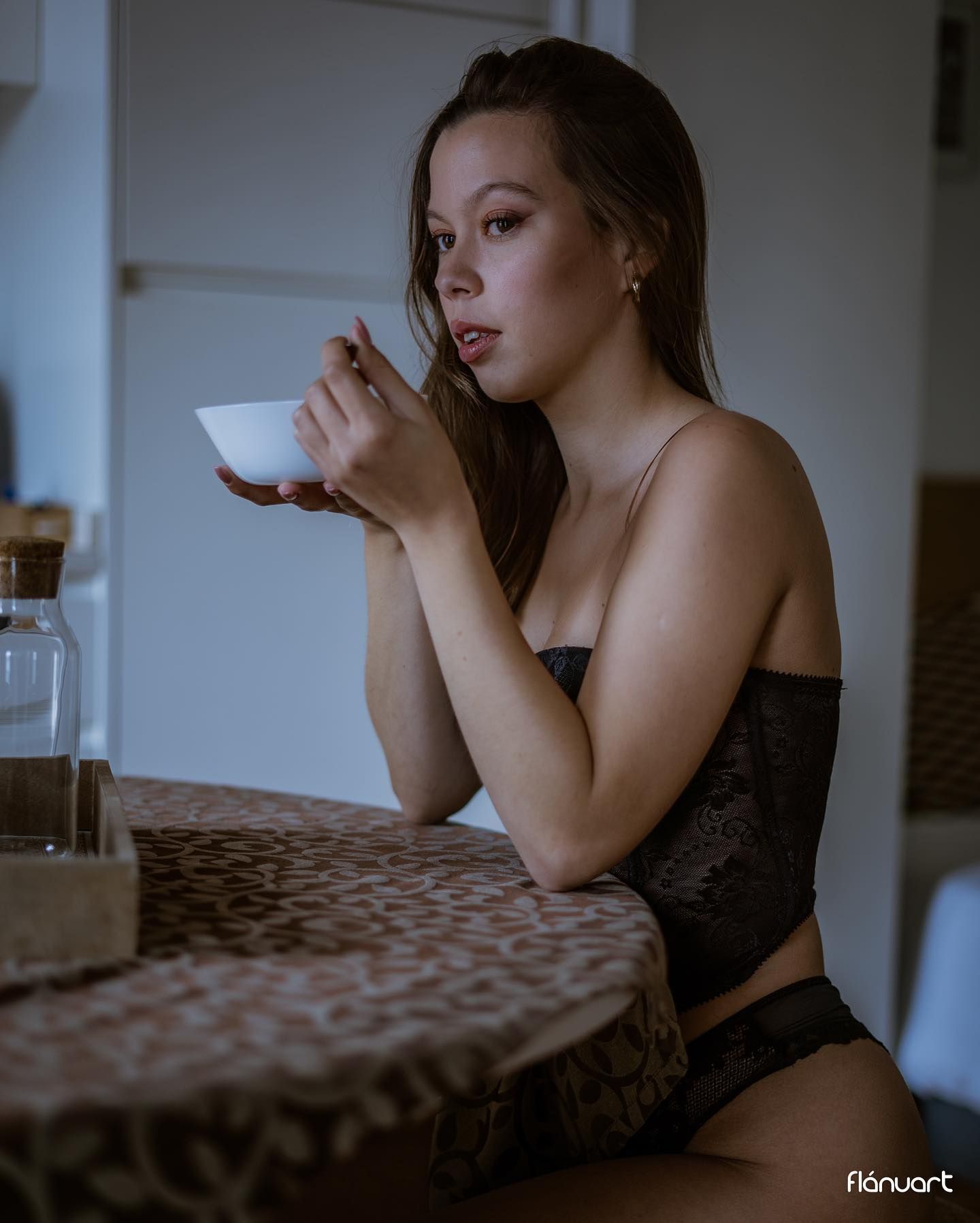 Brunette woman in black lace lingerie sipping from a bowl at a table in dim indoor lighting.