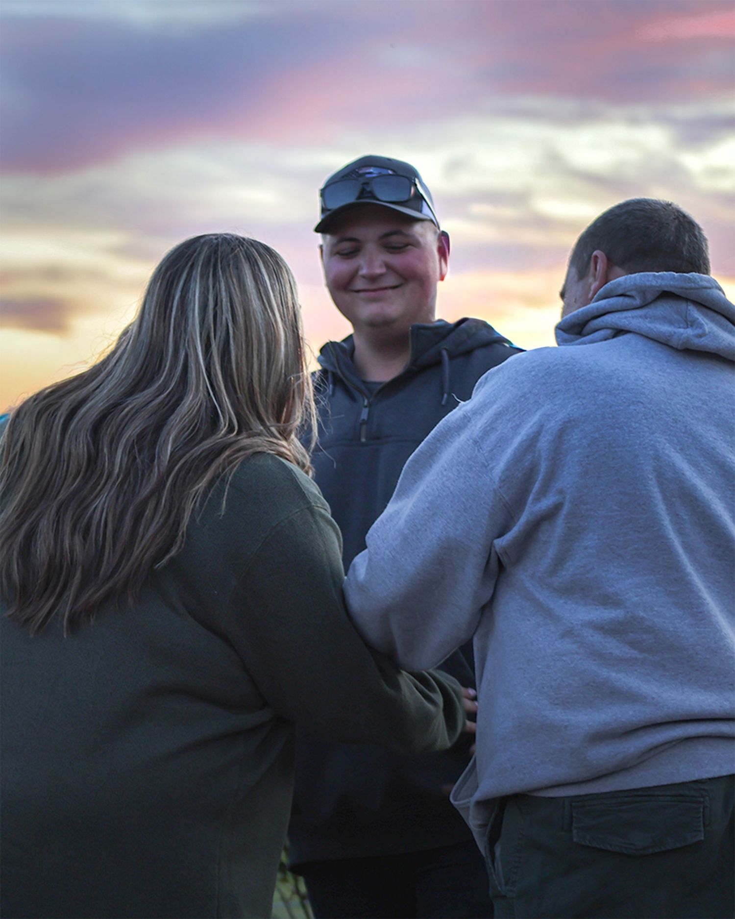 Young man smiling at sunset with two people facing away, sharing a warm outdoor moment with colorful sky.