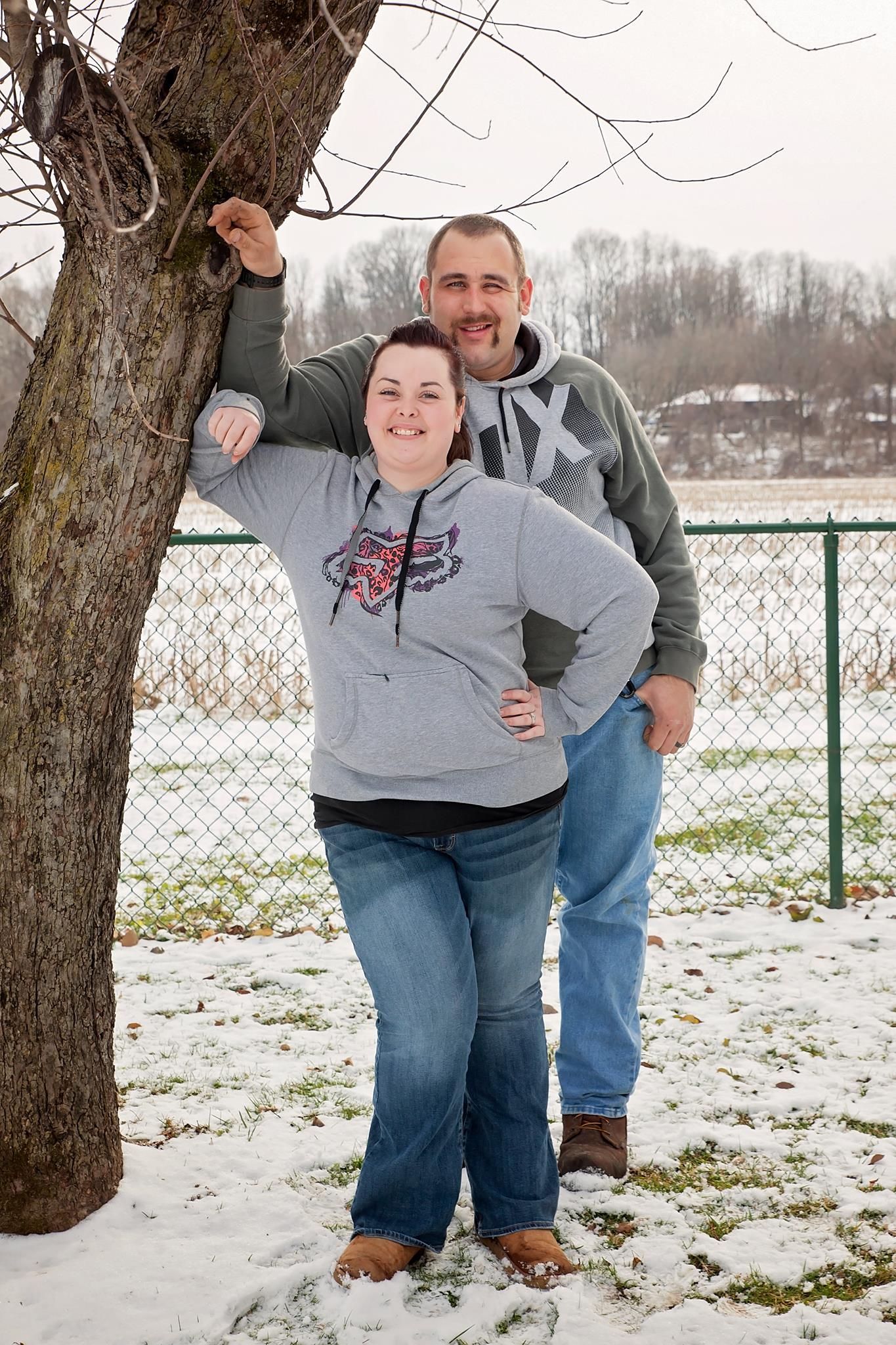 Smiling couple in hoodies posing by a bare tree in a snowy winter outdoor setting.