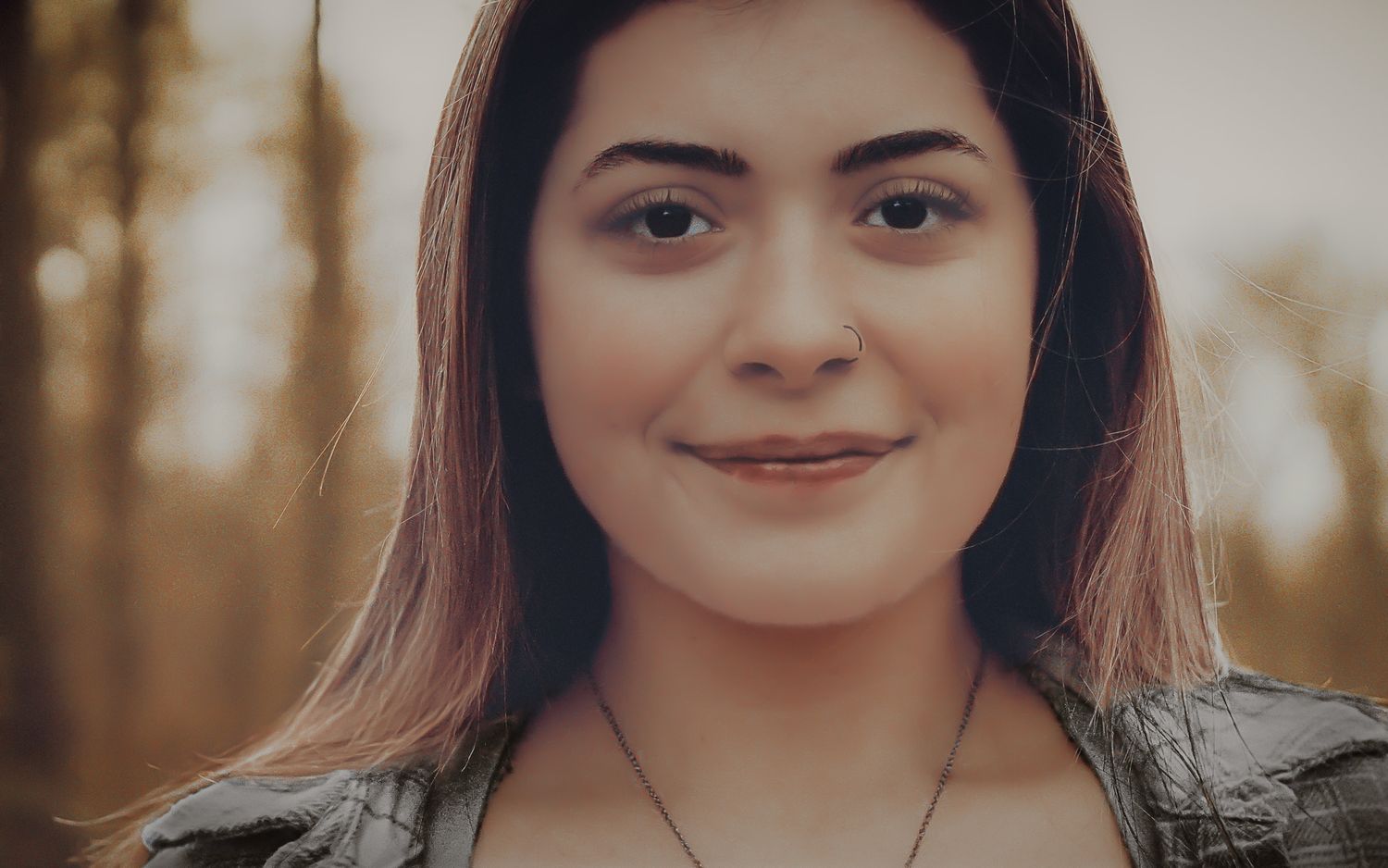 Young woman with long hair smiling softly outdoors in a warm bokeh forest setting.