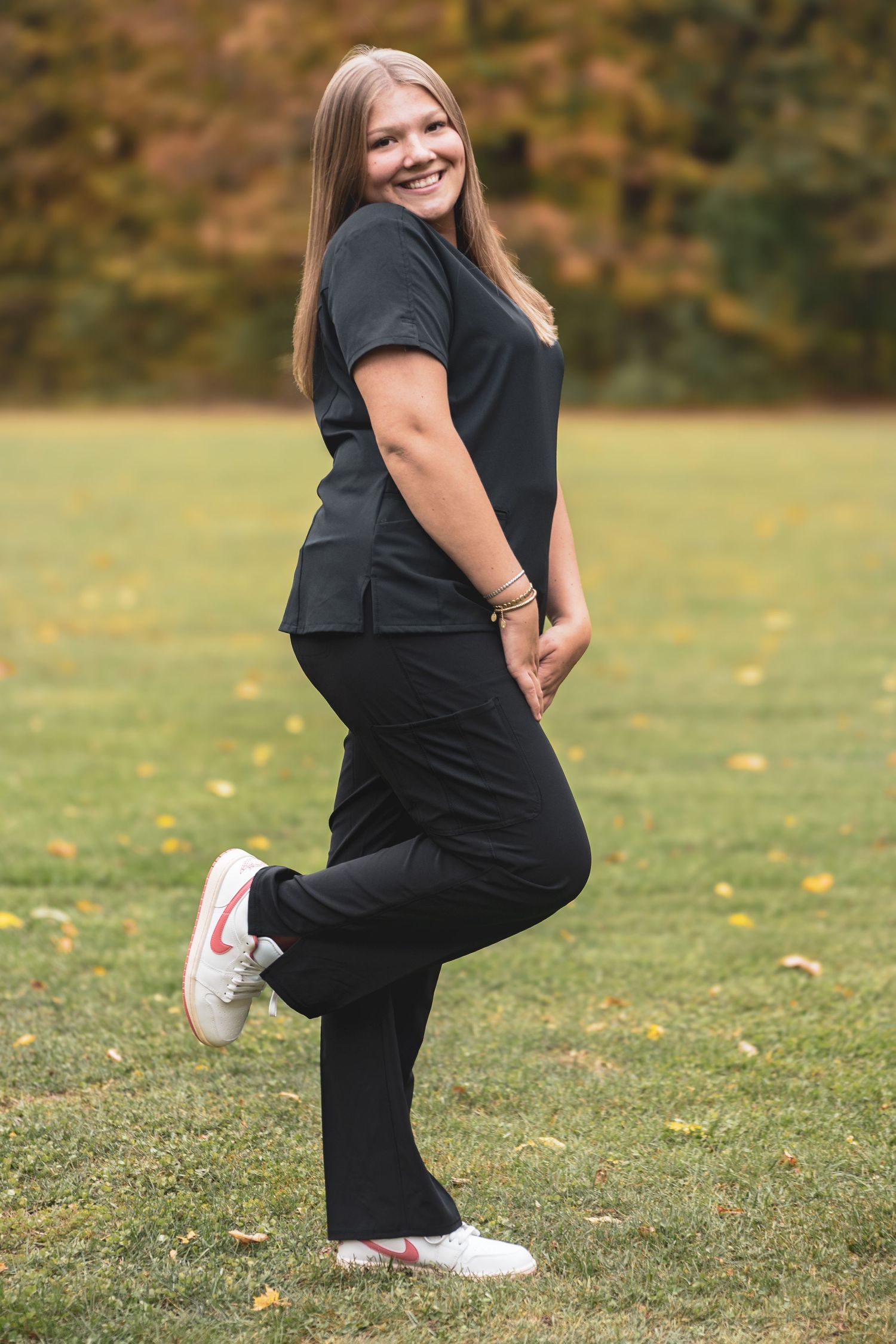 Smiling young woman in black outfit and white Nike sneakers posing in autumn park setting.