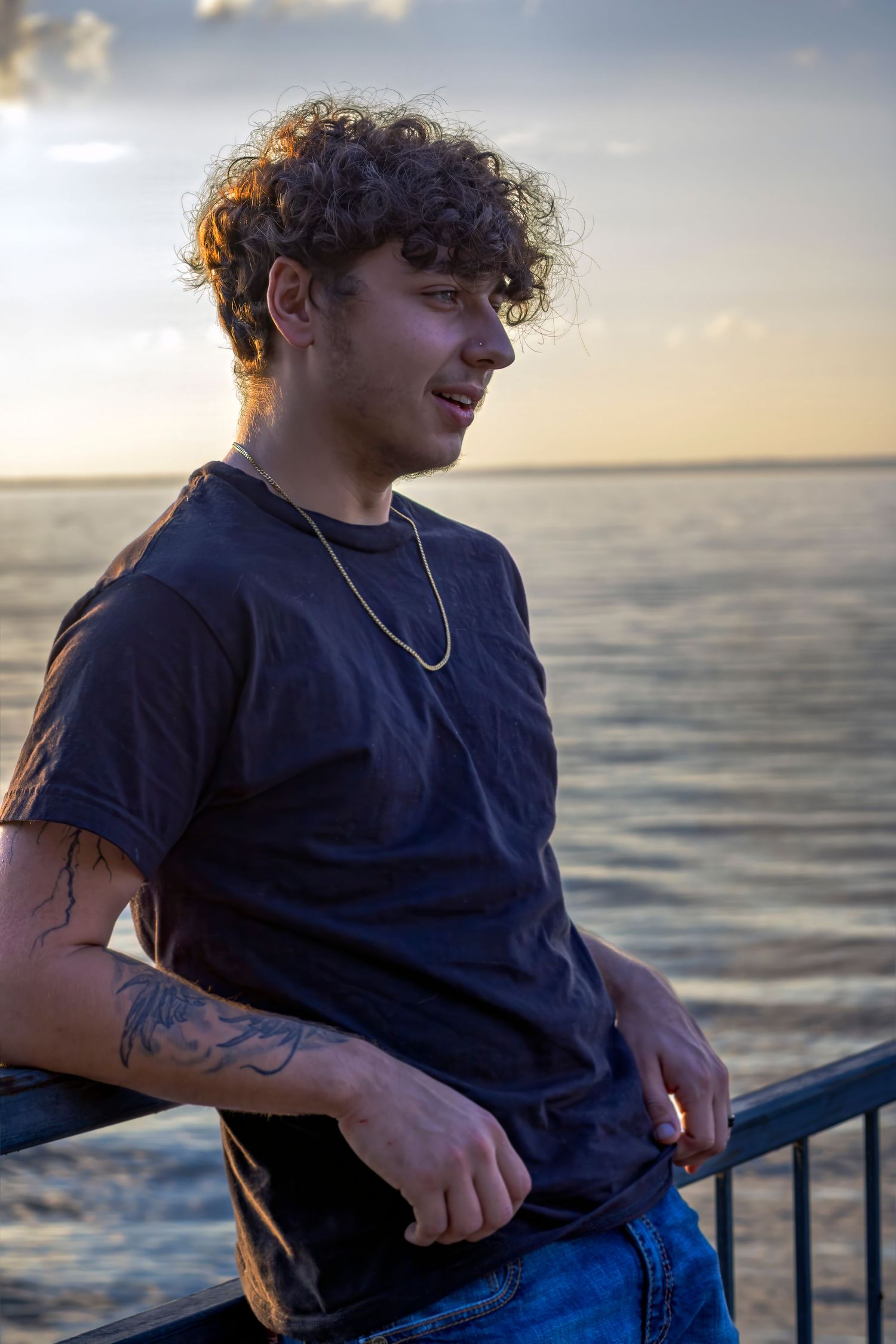 Young man with curly hair and arm tattoos in black t-shirt smiling on pier at golden hour sunset.