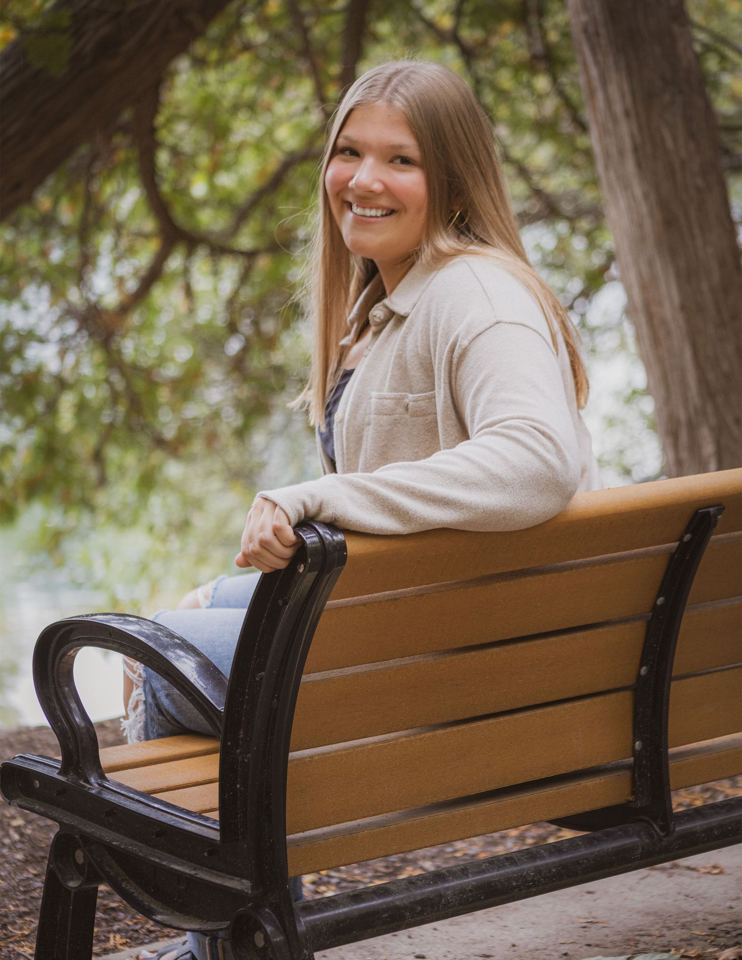 Young woman with long blonde hair smiling on a wooden park bench surrounded by trees.