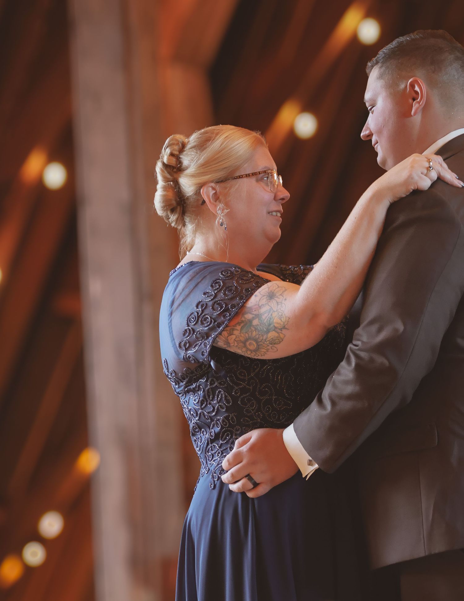 A woman with a tattoo in a navy lace dress dances with a man in a suit at a rustic barn wedding reception.