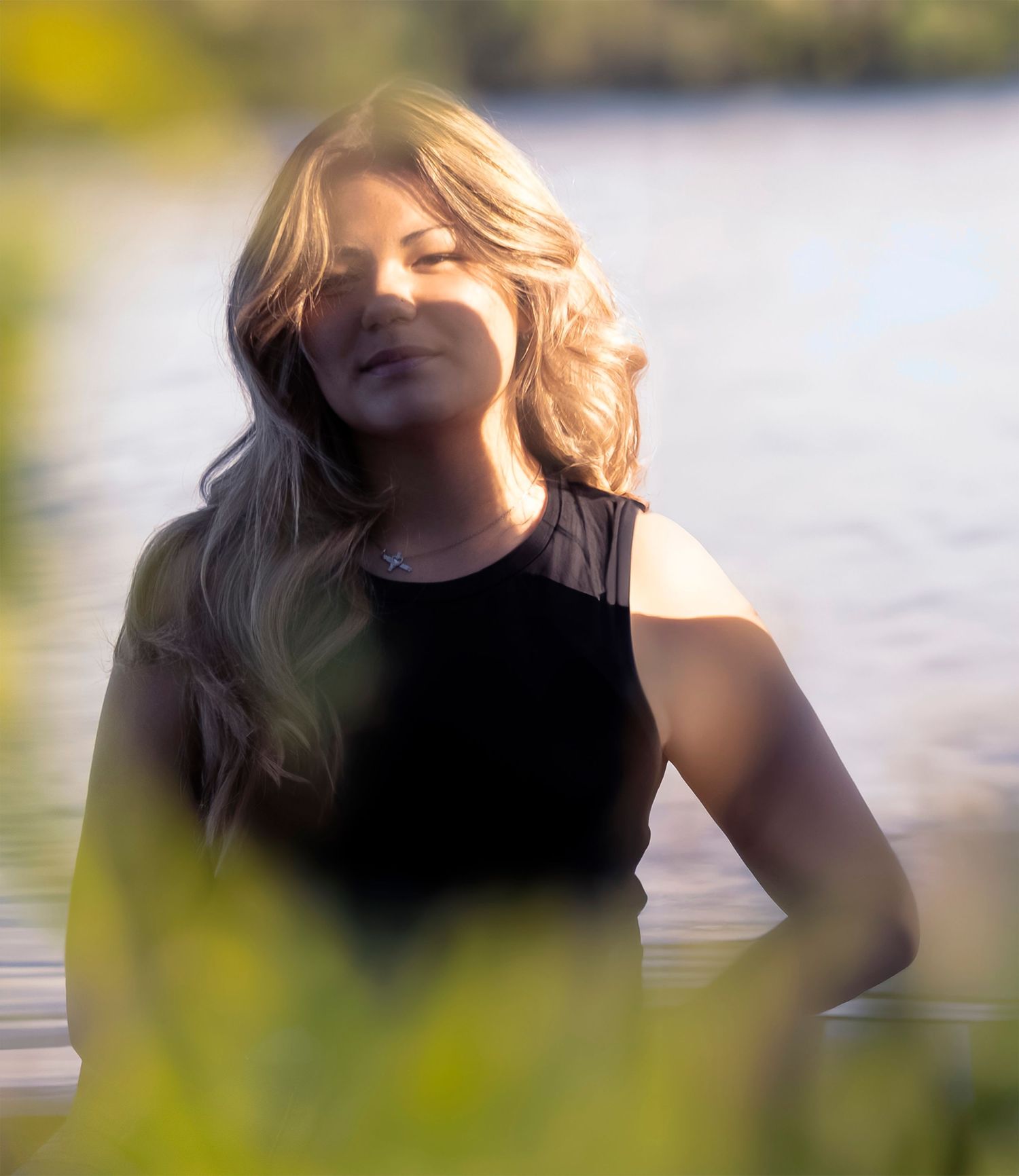 Young woman with blonde hair standing in a sunlit lake, eyes closed, wearing a black top.