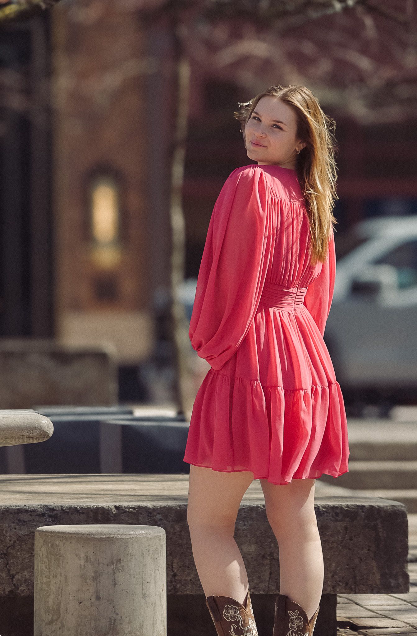 Young woman in coral pink mini dress and brown cowboy boots posing outdoors on a sunny day.