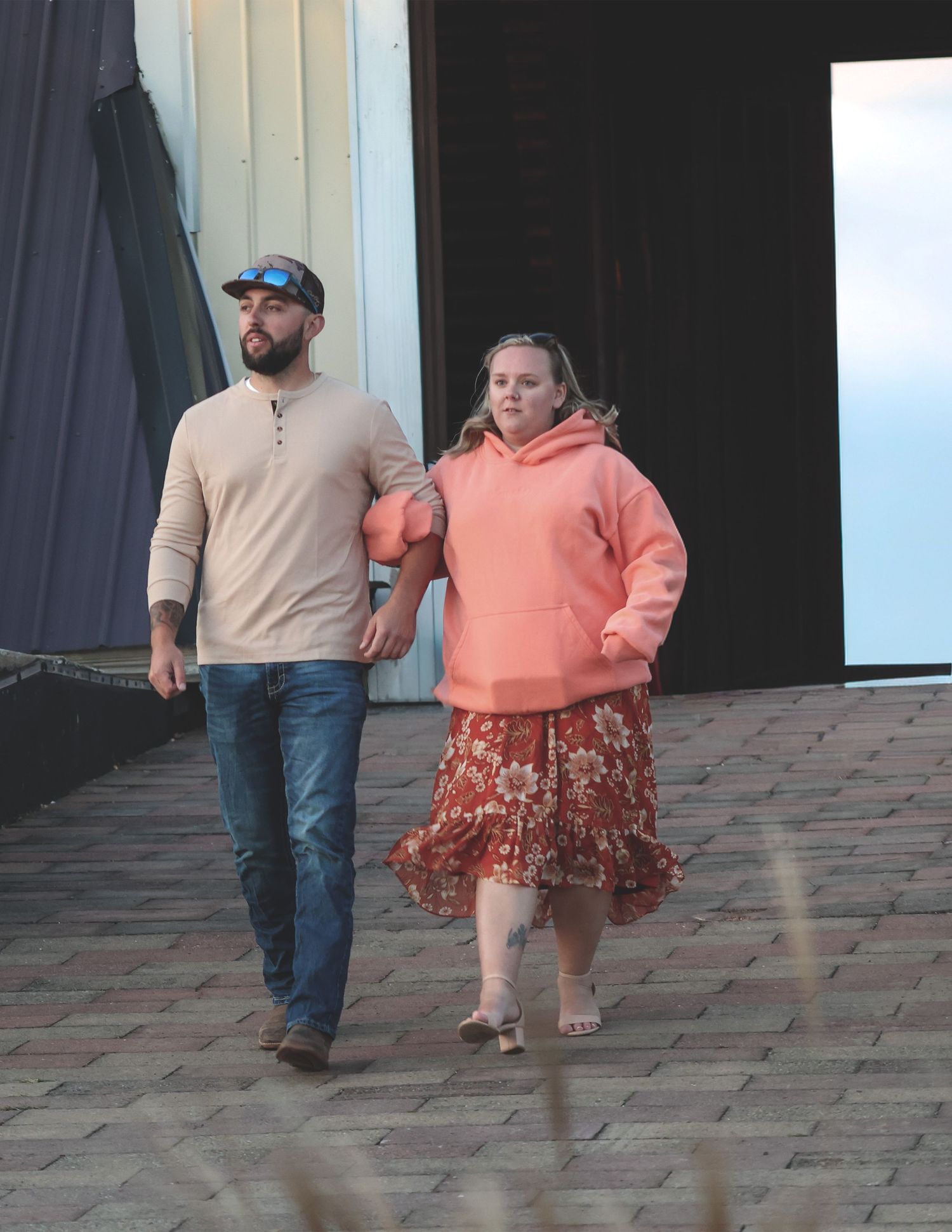 Couple walking arm in arm outdoors, man in beige shirt and jeans, woman in coral hoodie and floral skirt.