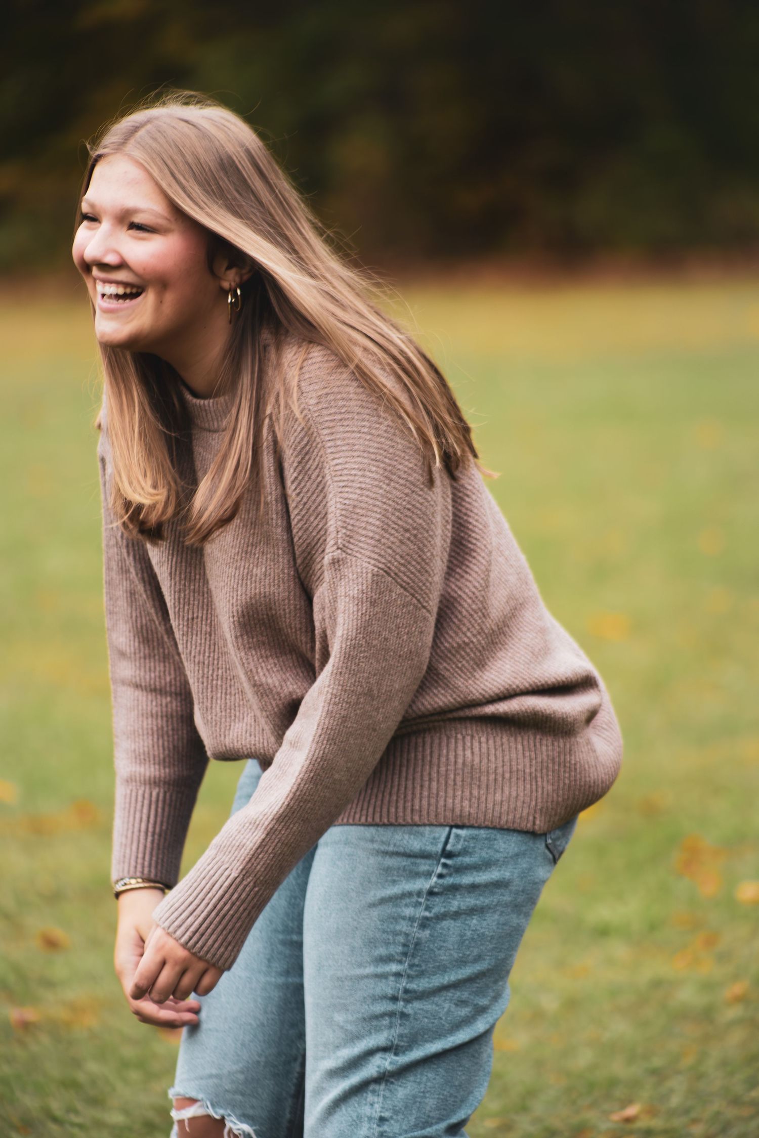 Laughing young woman with long blonde hair wears a brown knit sweater in an autumn outdoor setting.