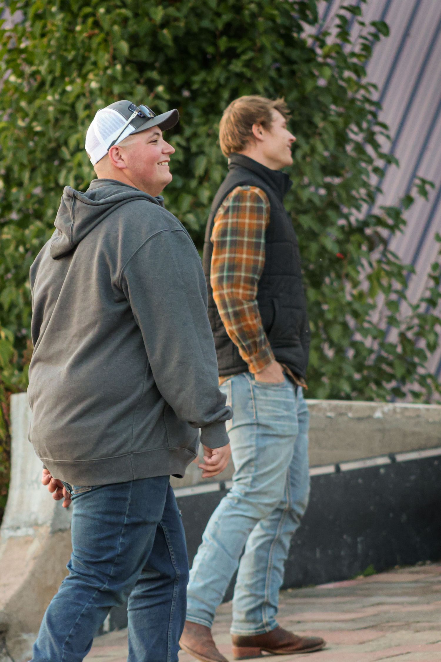 Two men in casual jackets and jeans walking outdoors near green foliage wall laughing together.