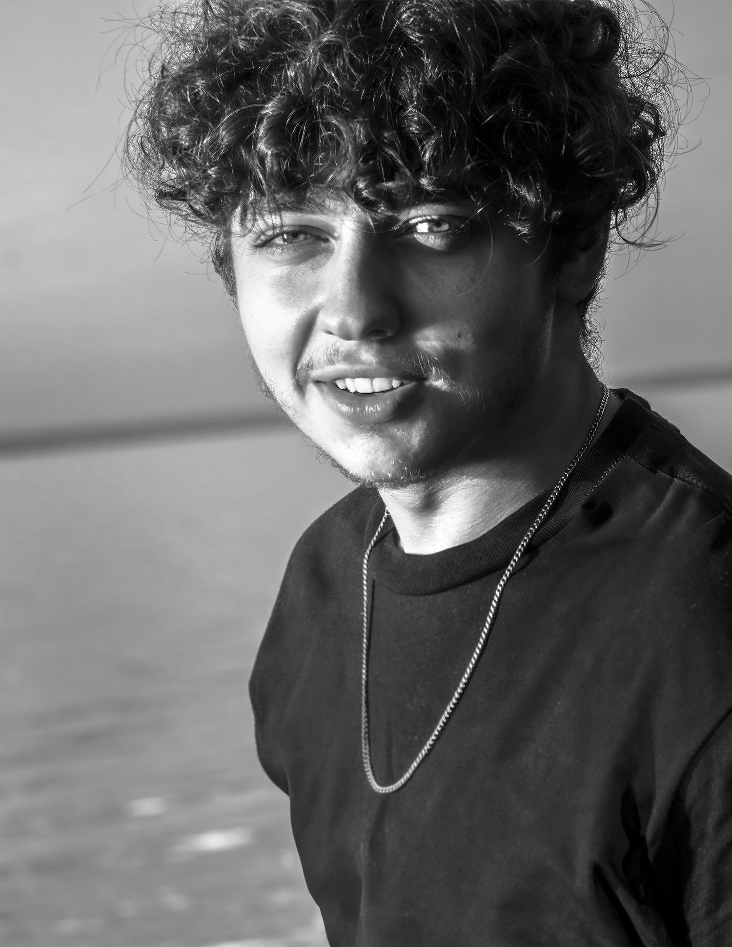 Black and white portrait of a young man with curly hair and chain necklace smiling outdoors near water.
