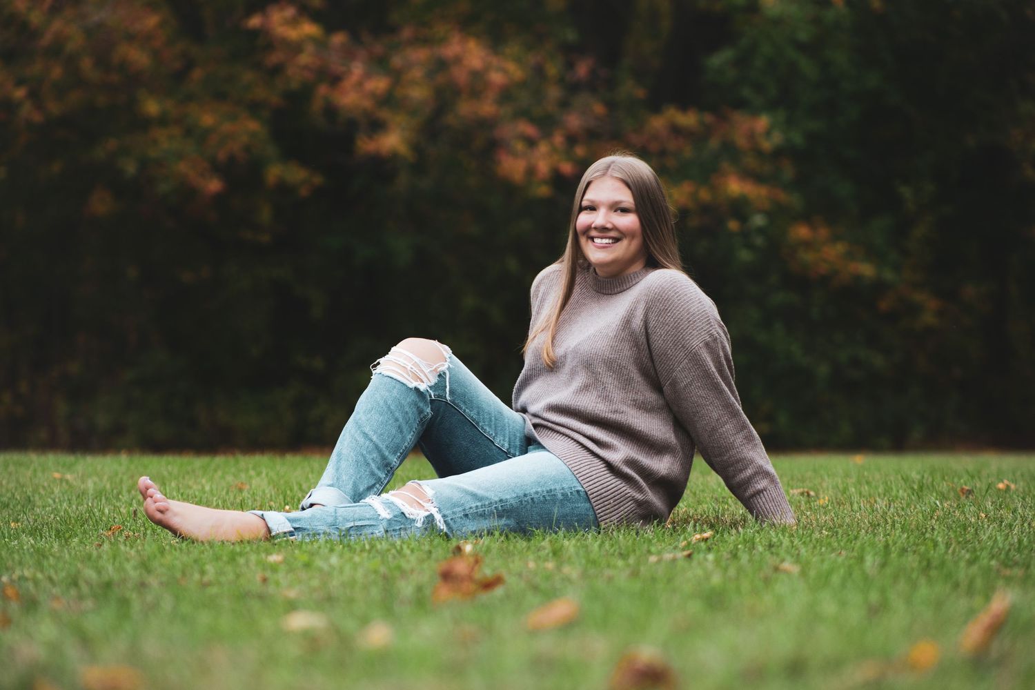 Young woman smiling in ripped jeans and gray sweater sitting on green grass with autumn trees in background.