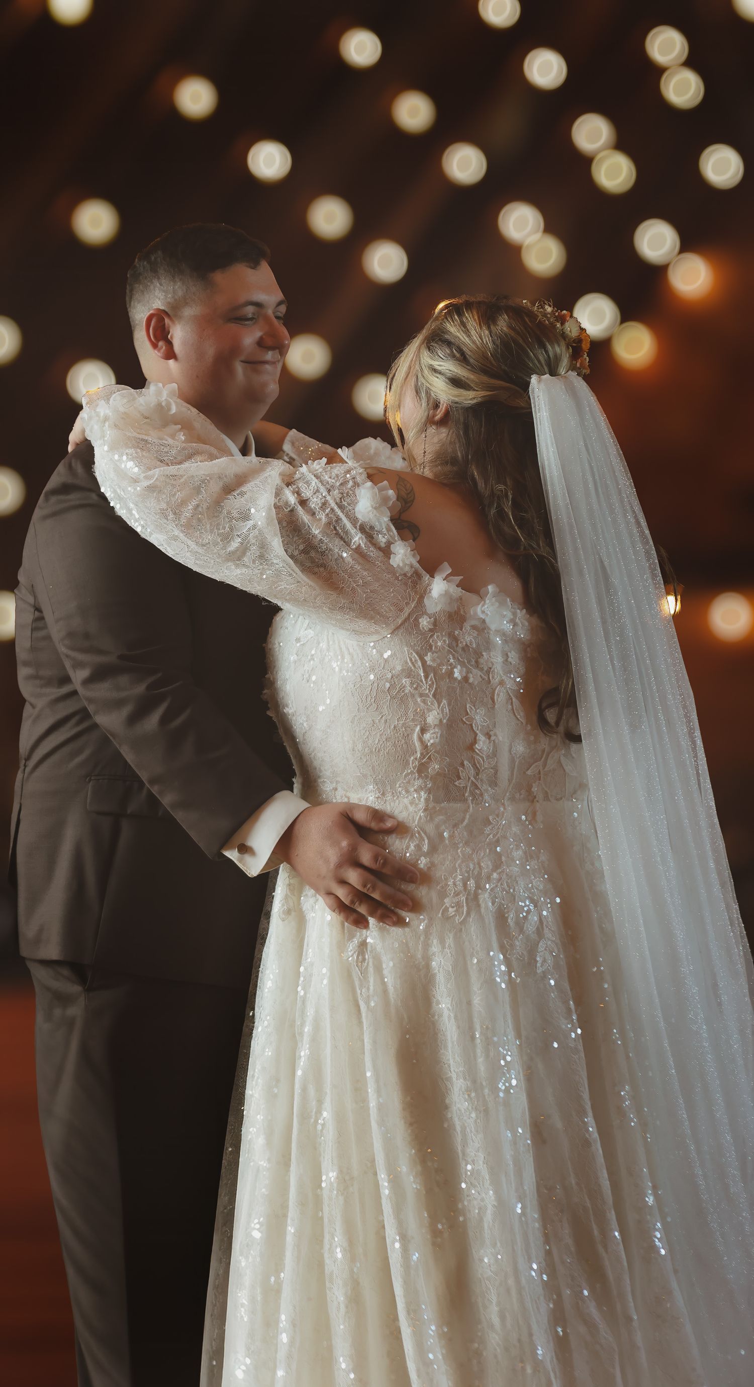 Bride and groom share first dance under warm bokeh string lights in rustic barn wedding venue.