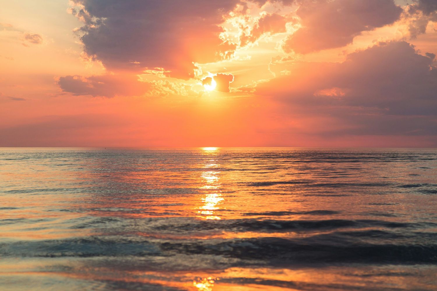 A stunning ocean sunset with vibrant orange and red hues reflecting on calm water beneath dramatic clouds.