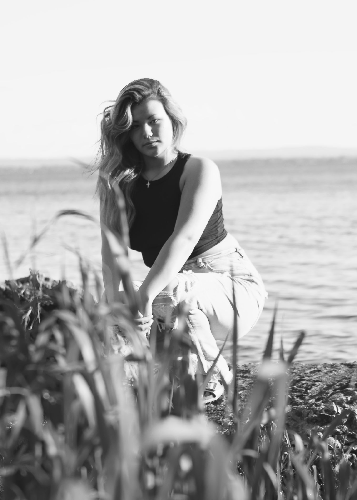 Black and white photo of a woman crouching near tall grass by the water's edge.