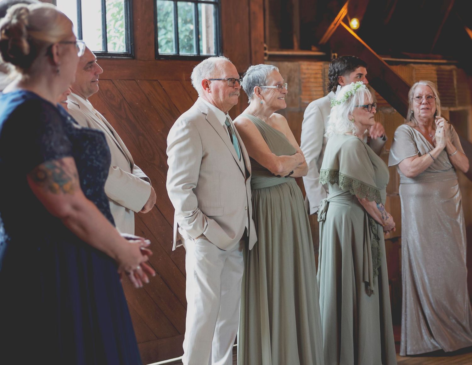 Wedding guests in formal attire stand watching a ceremony inside a rustic wooden barn venue.