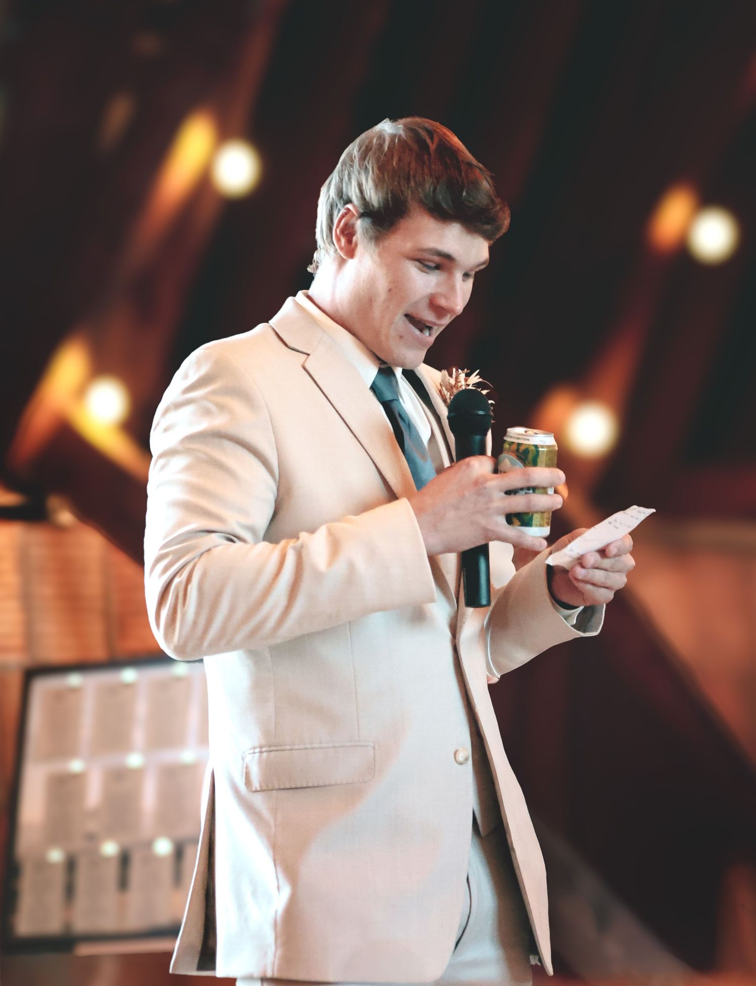 A man in a cream suit speaks into a microphone while reading from a card at a formal event.