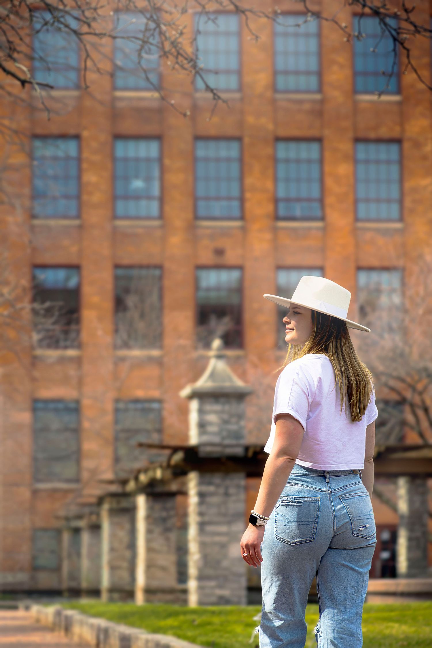 Young woman in white hat and jeans poses outdoors in front of a red brick building on a sunny day.