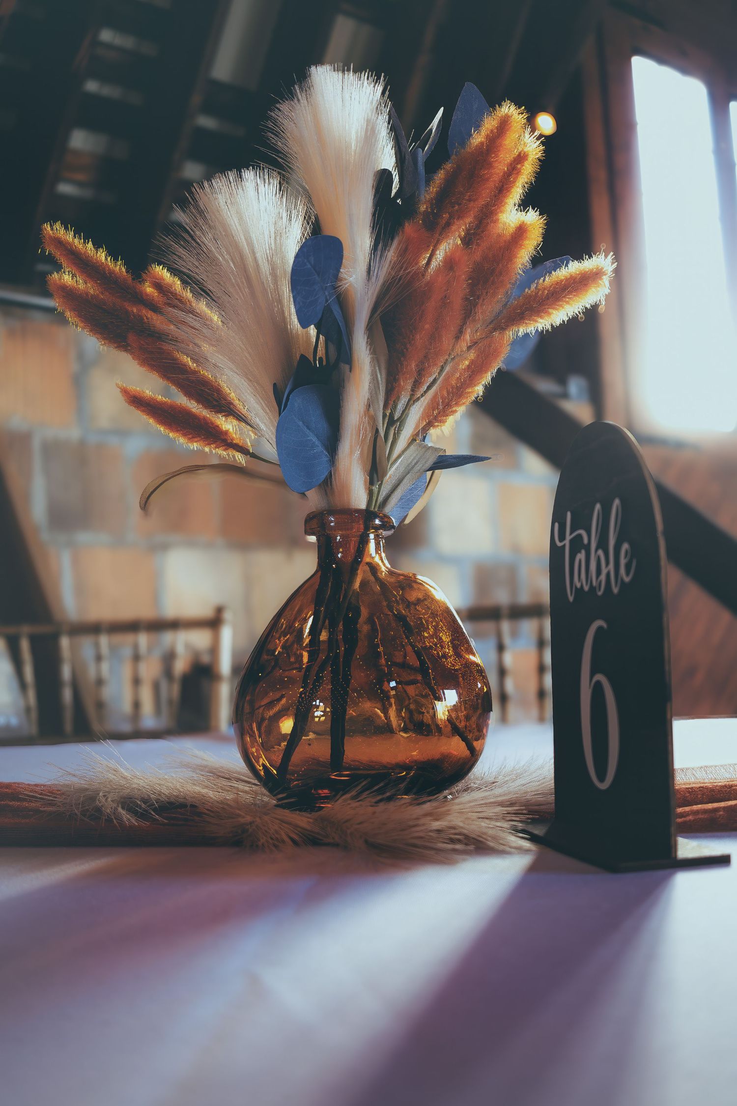 Rustic wedding table centerpiece with dried pampas grass and blue feathers in amber vase beside Table 6 sign.