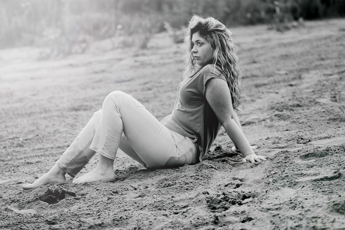 Black and white portrait of a woman sitting on sandy ground outdoors in casual attire.