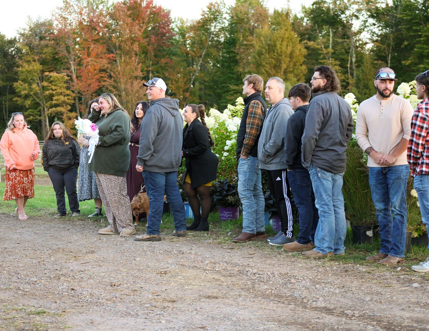 Group of people standing in line outdoors near trees and shrubs on a gravel path during autumn.