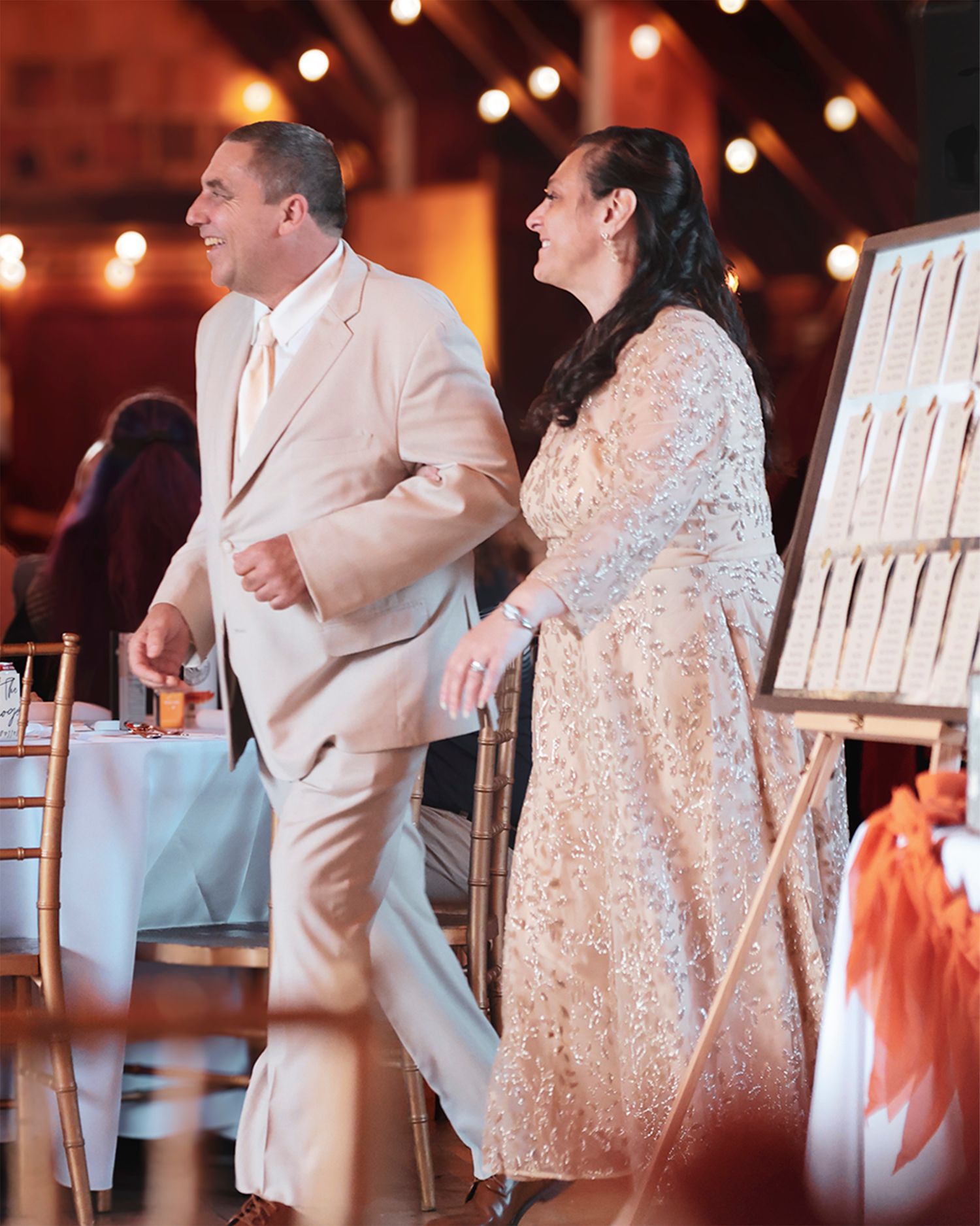 Couple in elegant attire smile as they enter a warmly lit wedding reception venue.