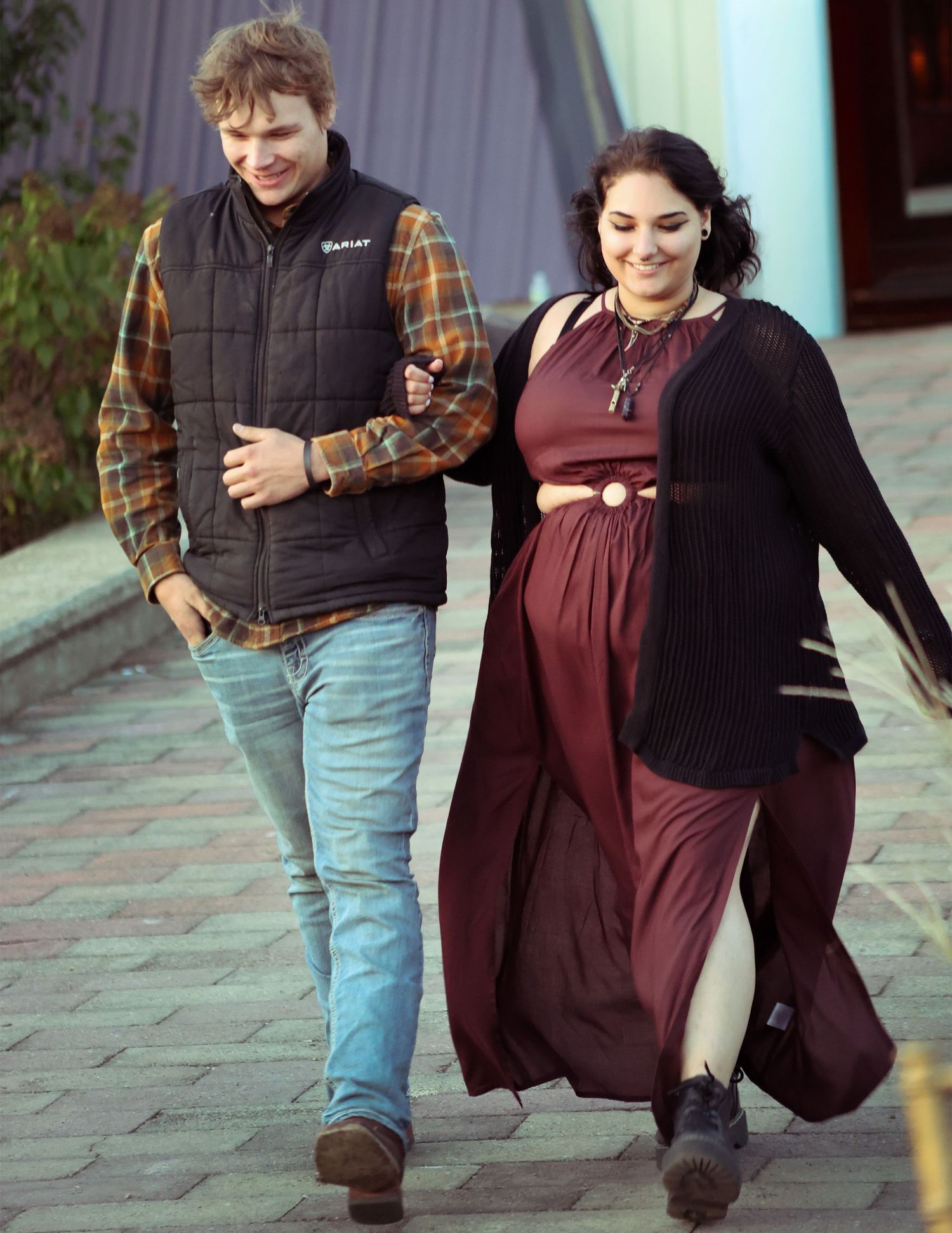 A couple walks arm in arm outdoors, man in plaid shirt and vest, woman in burgundy maxi dress and black cardigan.