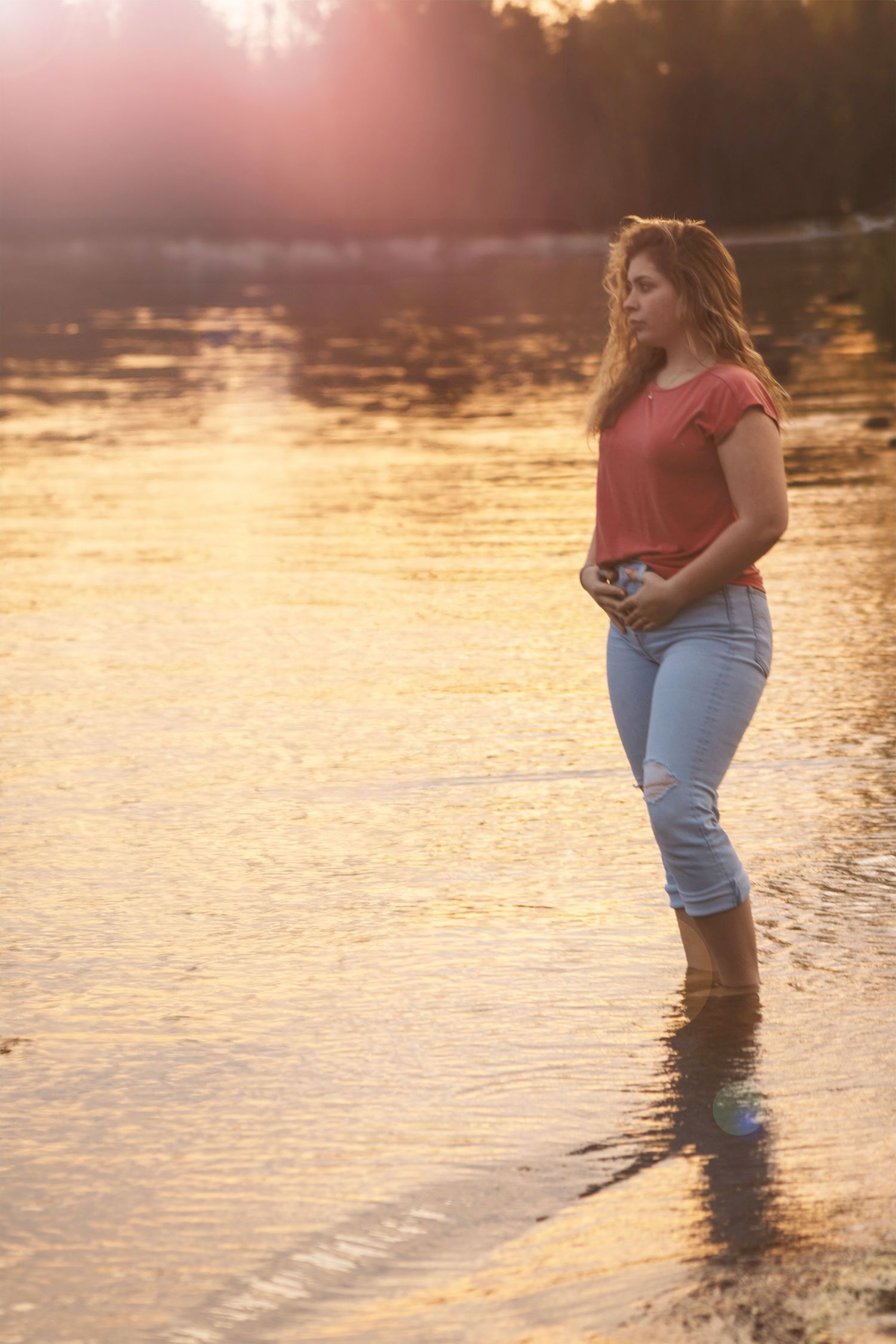 Young woman in jeans and pink top standing in golden sunset water, serene nature scene.