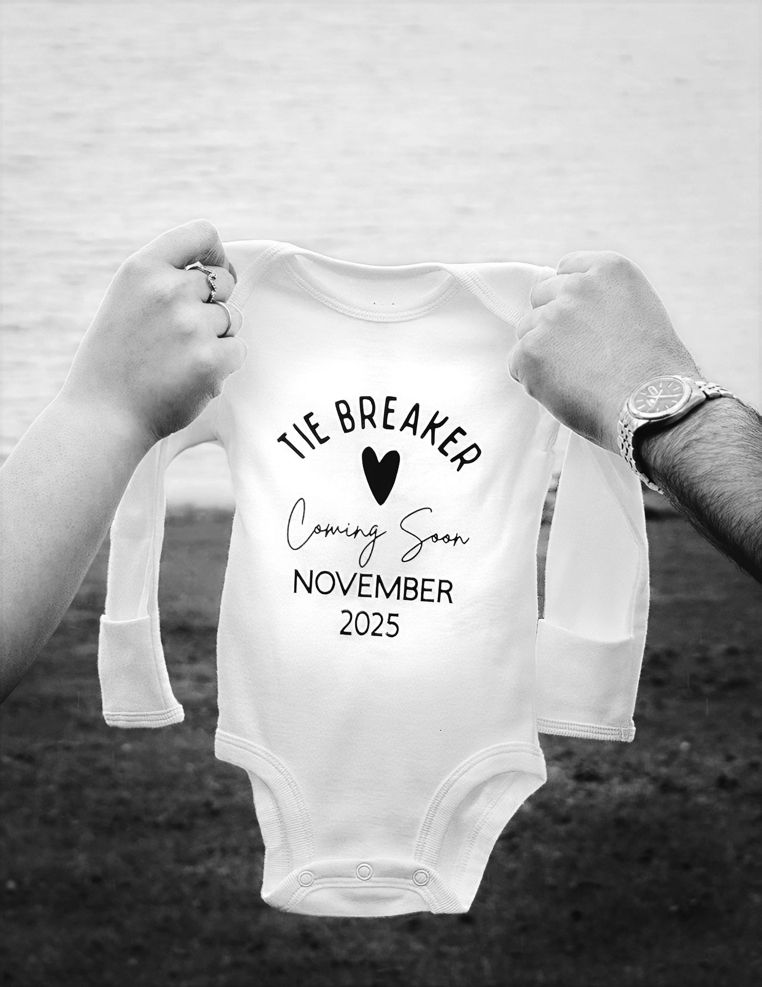 Hands holding a white baby onesie reading Tie Breaker Coming Soon November 2025.