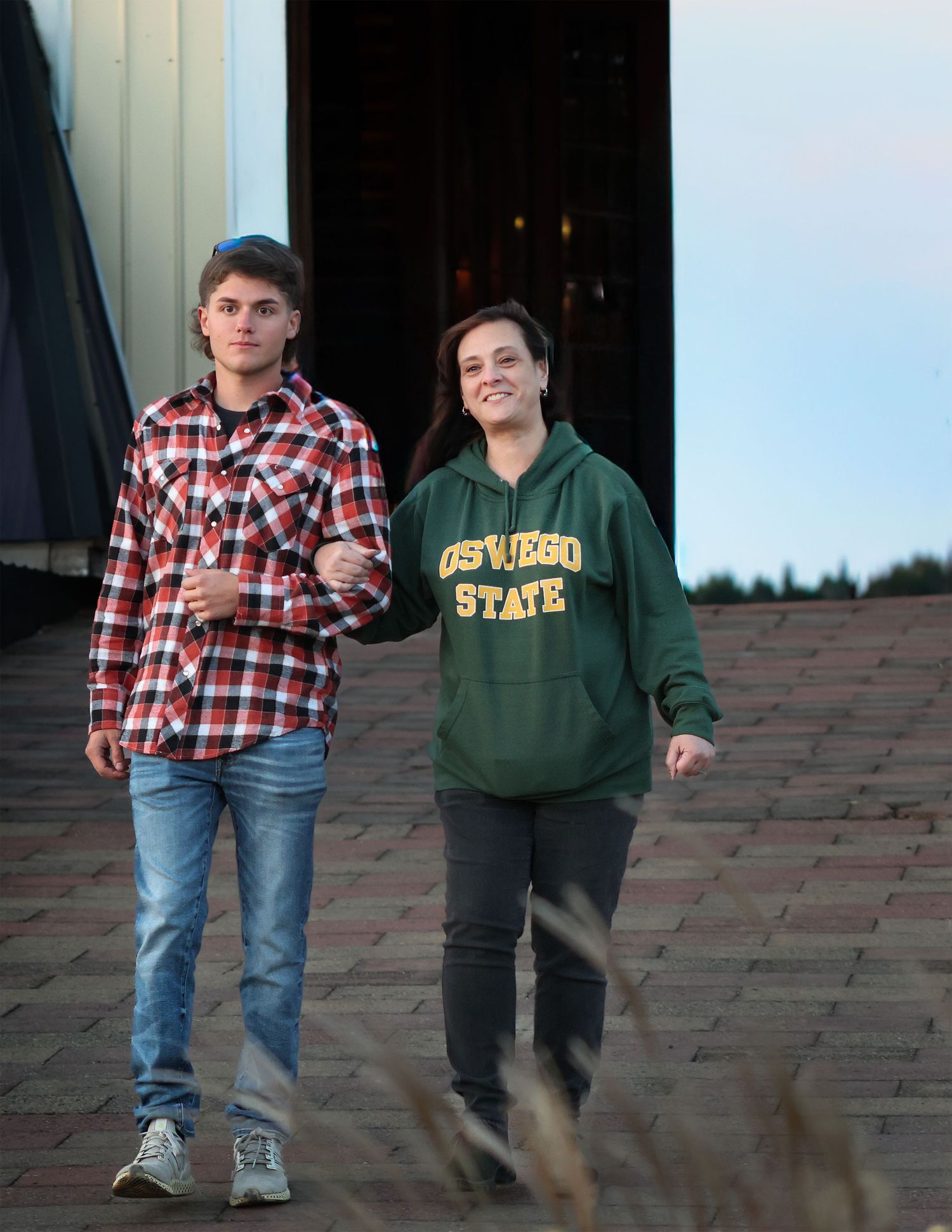 Two young people walking out of a barn, one wearing a plaid shirt and jeans, the other in an Oswego State green hoodie.