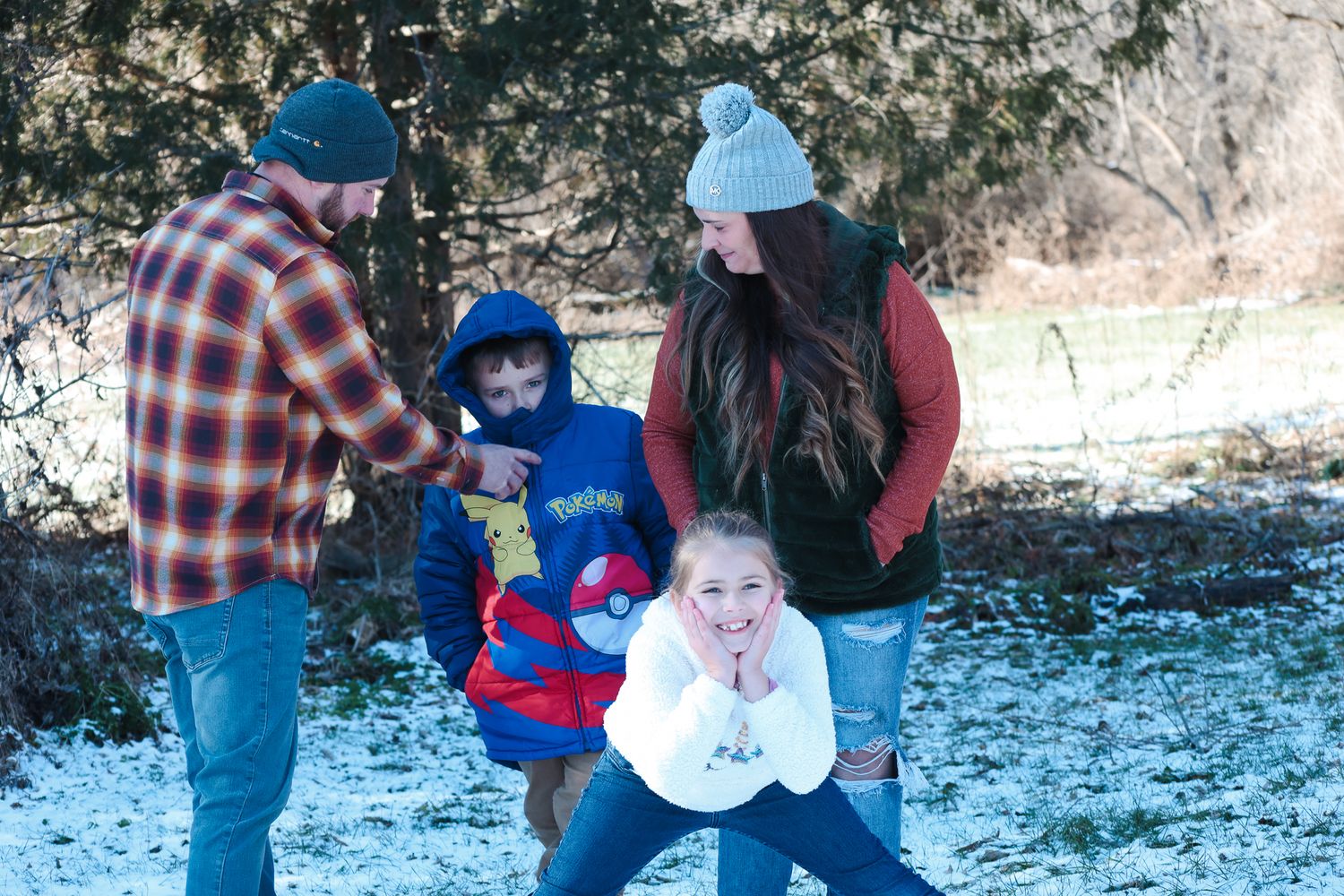 Family of four playing outdoors in light snow, wearing winter clothes among snow-dusted trees.