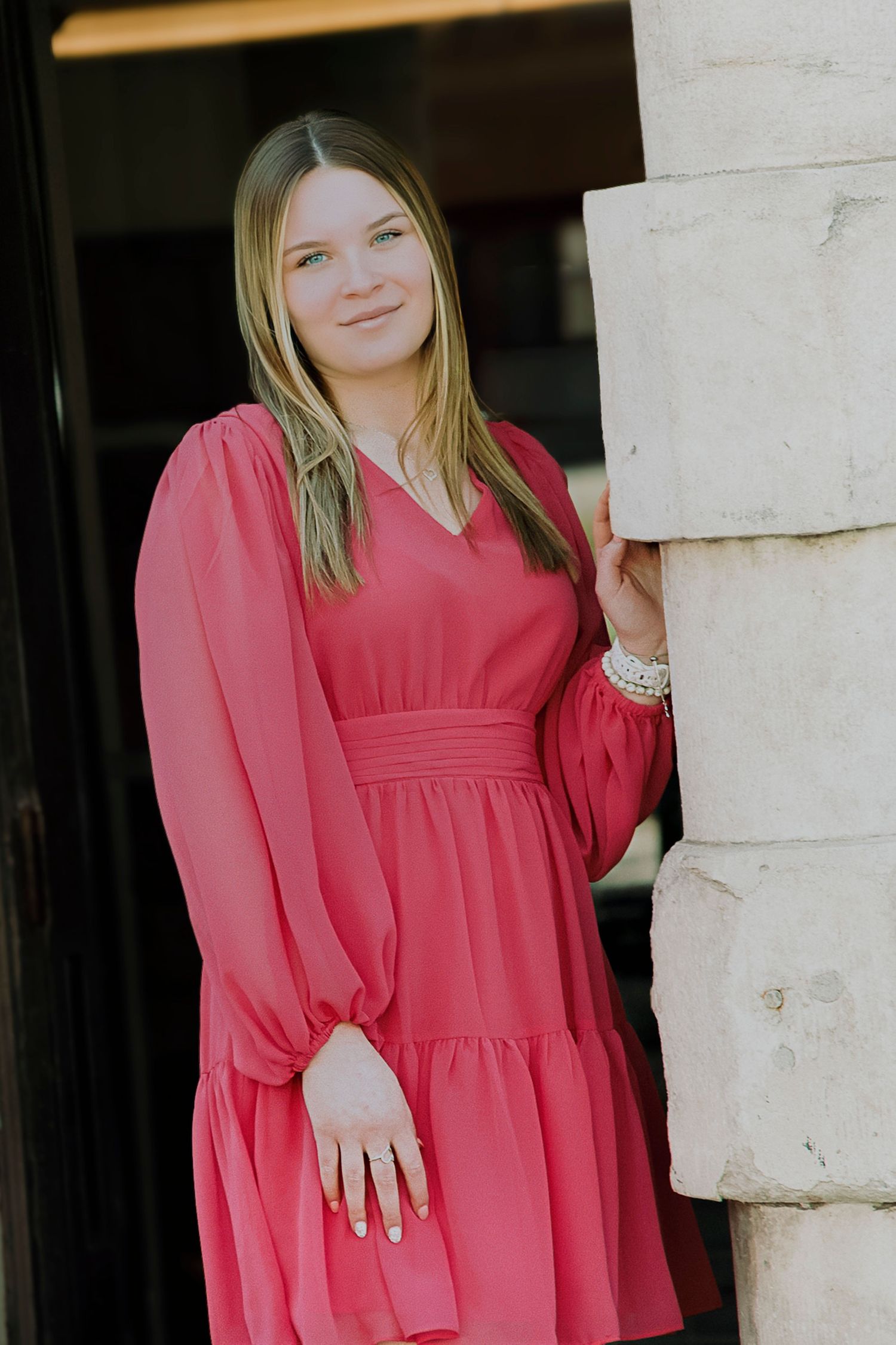 Young woman in a pink long-sleeve dress leaning against a stone column outdoors.
